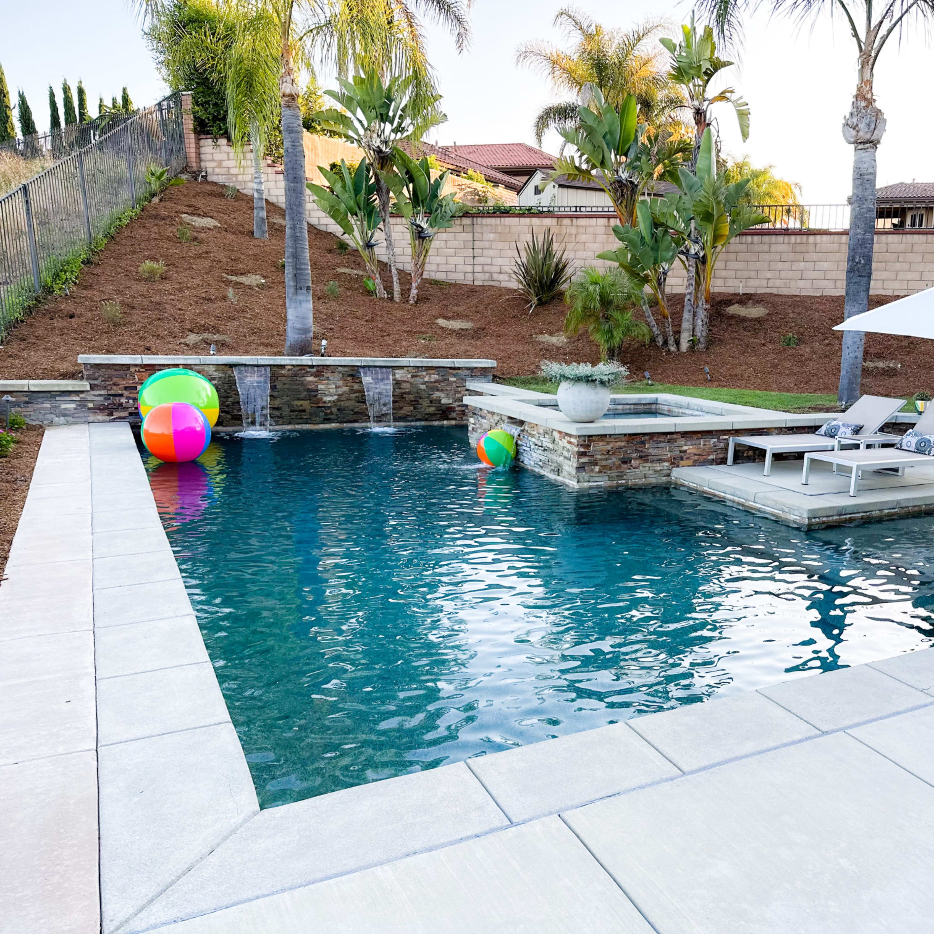 A swimming pool surrounded by palm trees features lounge chairs, a small hot tub, and colorful beach balls.