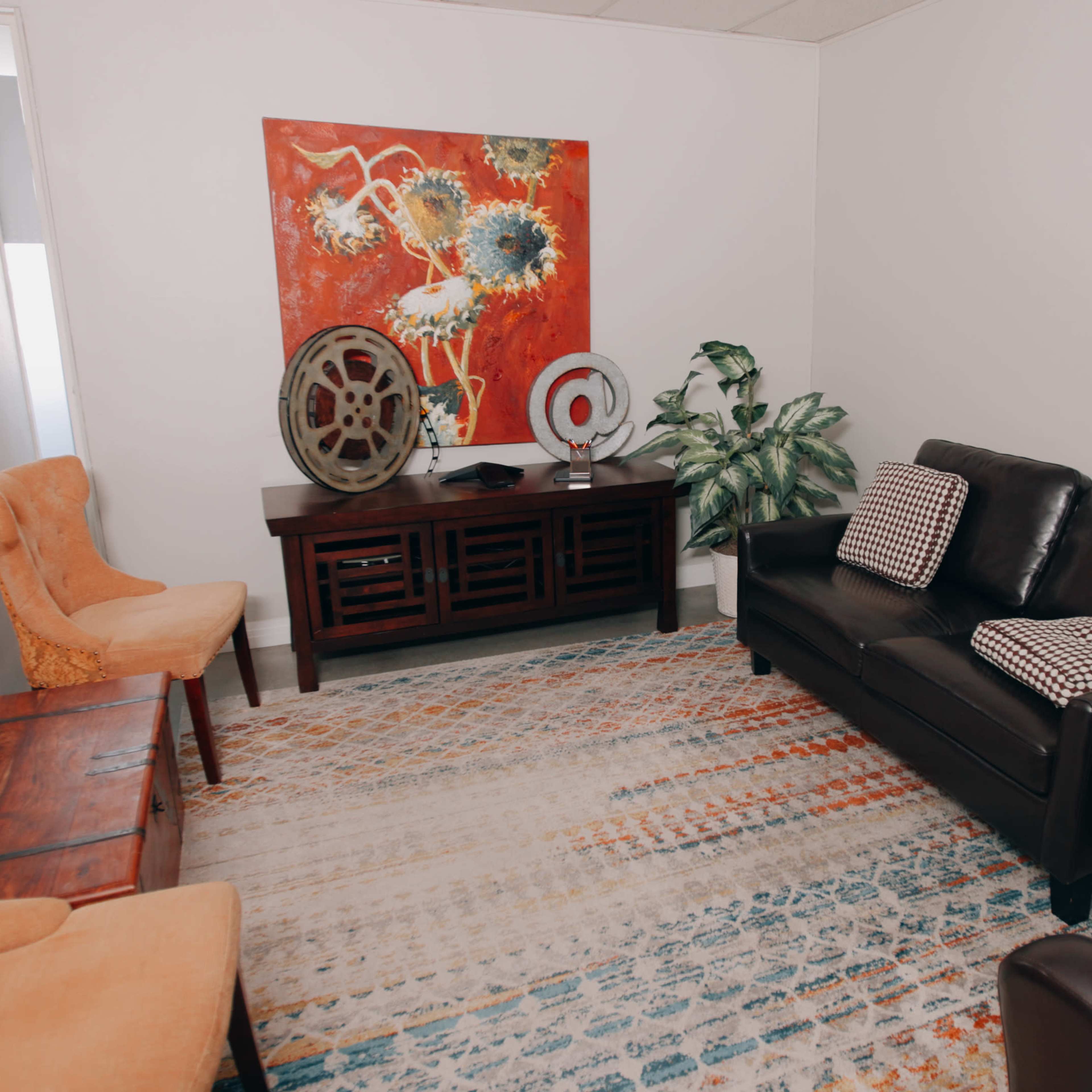A small, furnished room with a black sofa, two orange chairs, a wooden table, a colorful rug, and a large decorative painting on the wall.
