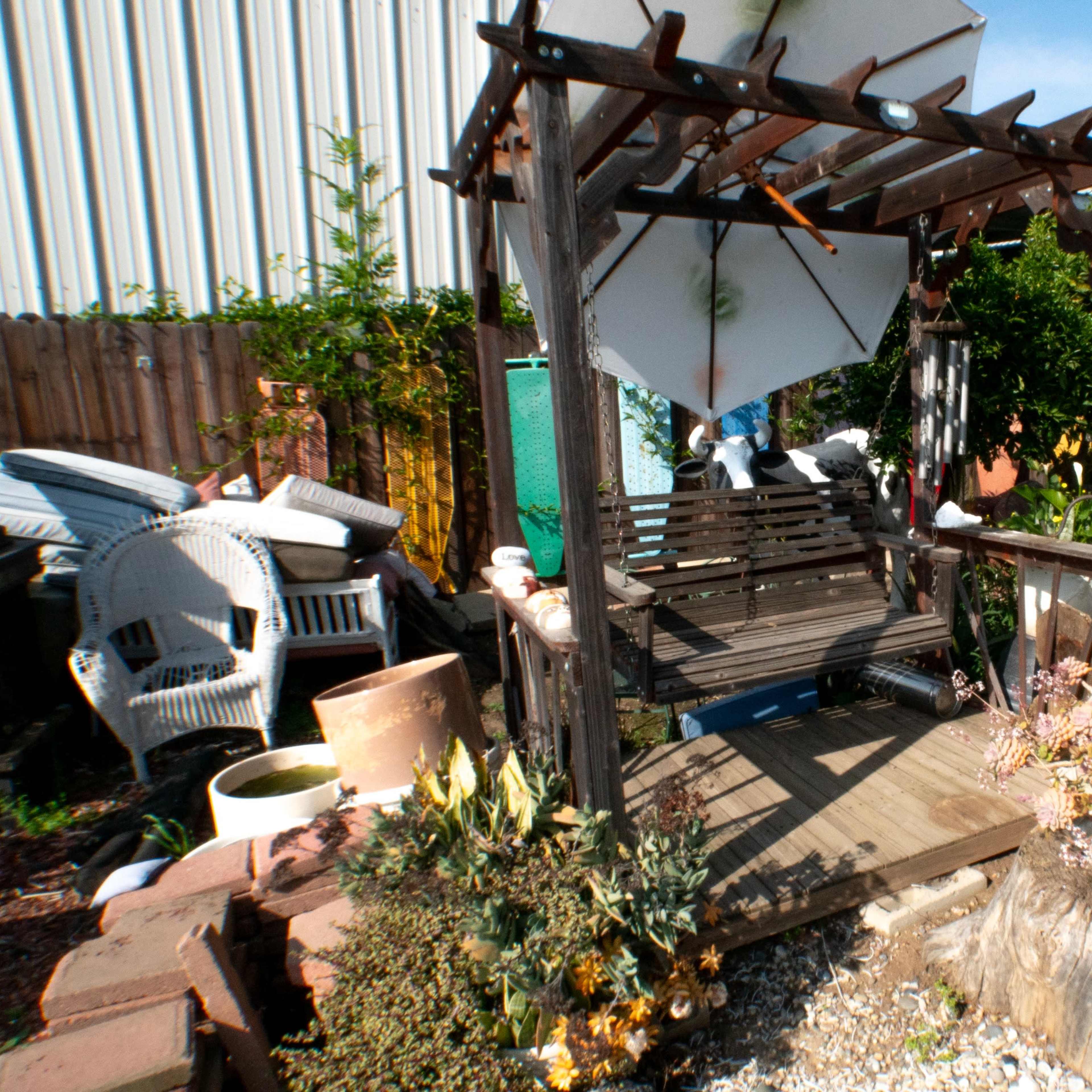 A wooden swing under a trellis surrounded by various plants and garden items in a cluttered backyard.