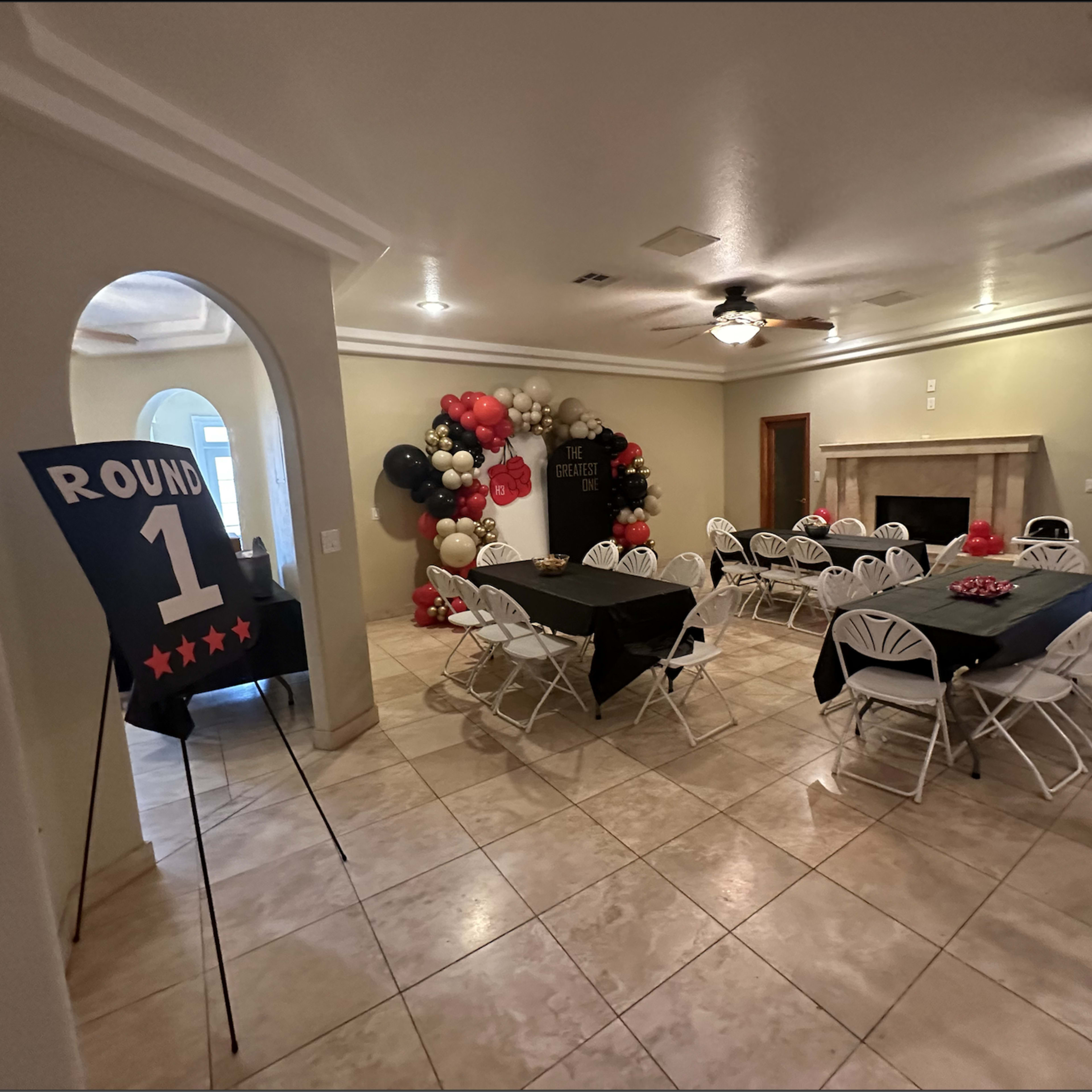 A spacious room is set up for an event, featuring tables covered with black cloths, white chairs, a large sign indicating "ROUND 1," and a decorative arrangement of balloons against the wall.