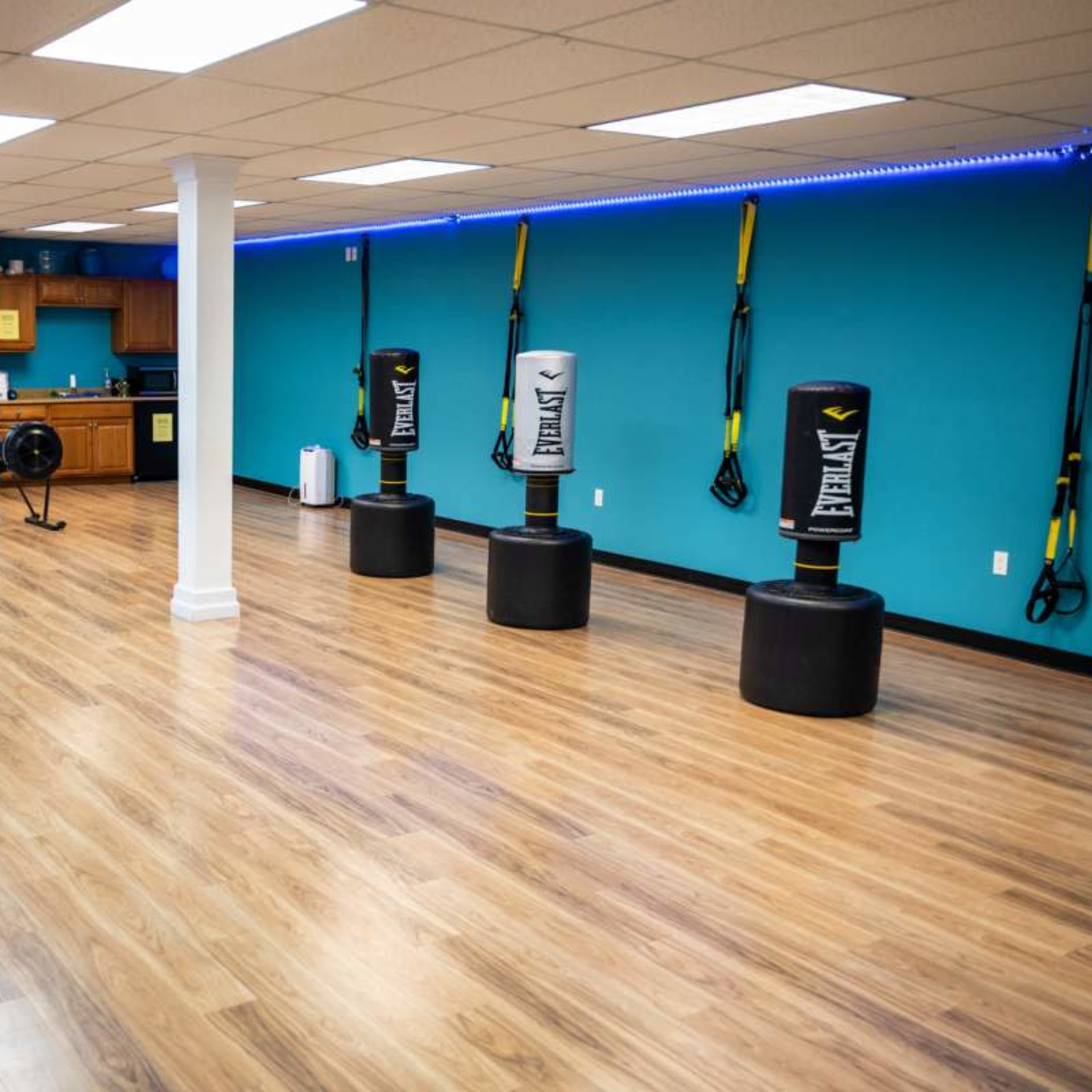 A fitness studio featuring three heavy punching bags, exercise equipment, and a kitchen area, all set against a blue accent wall.