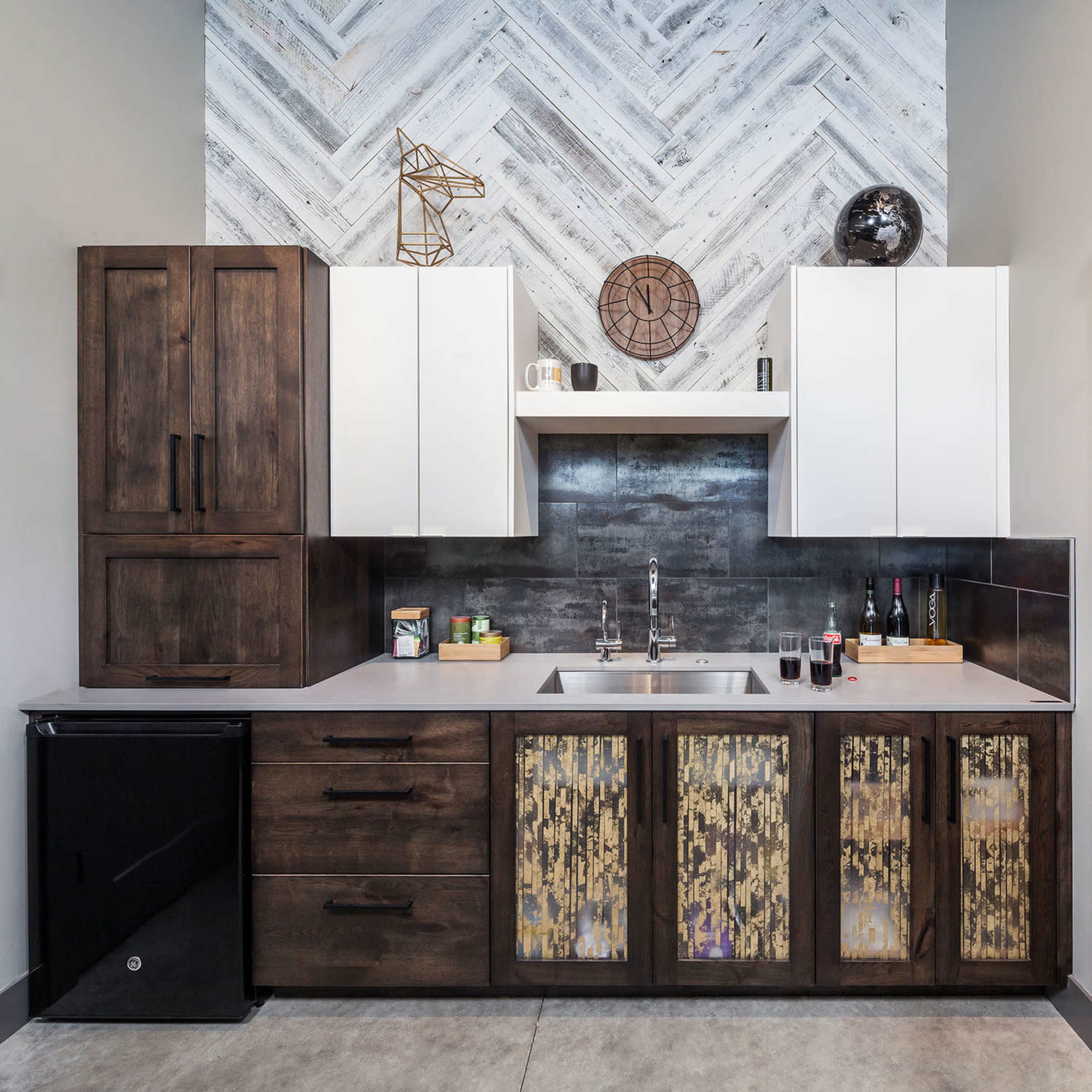 A modern kitchen with a herringbone-patterned accent wall, featuring dark wood cabinets, white upper cabinets, and a stainless steel sink with various kitchen items arranged on the countertop.