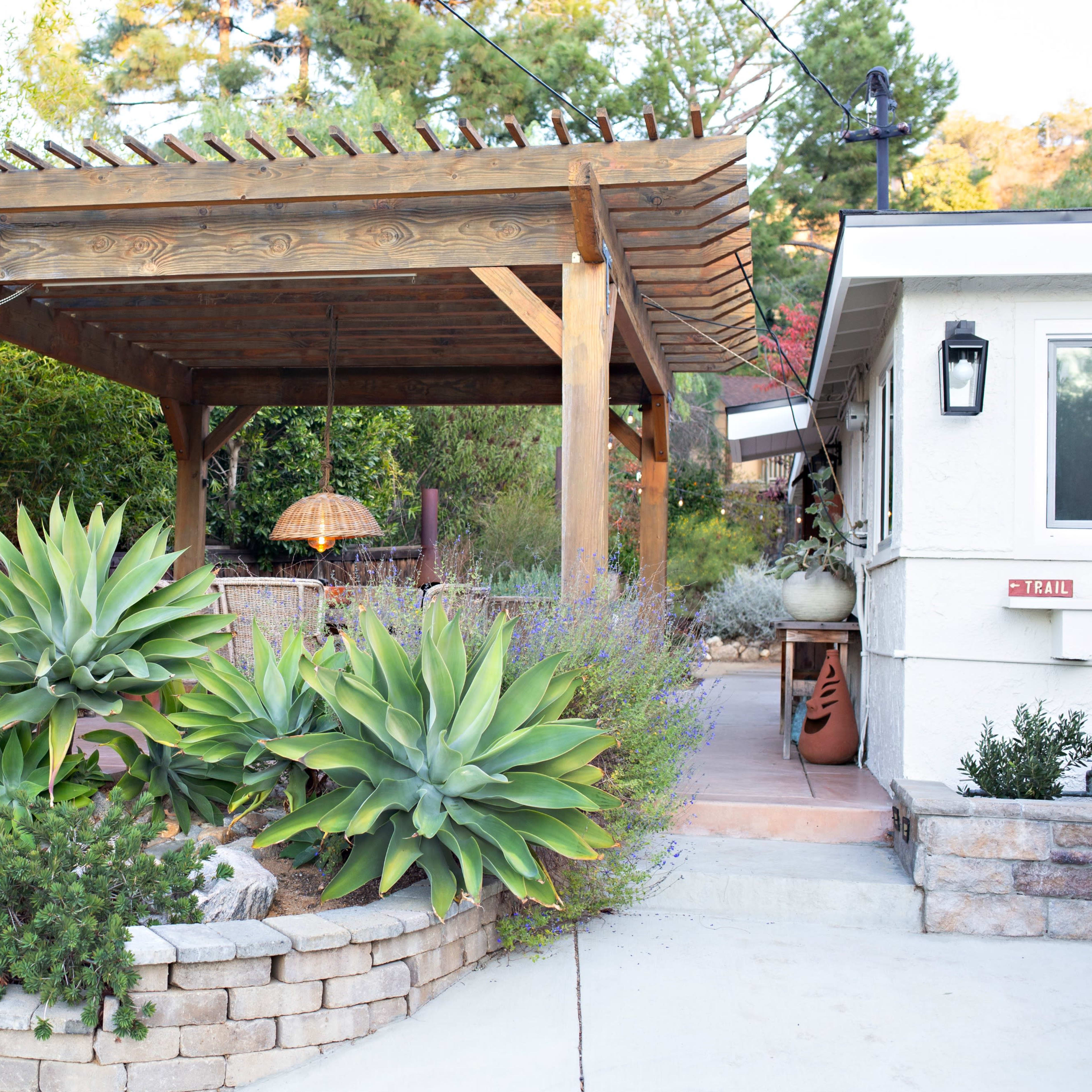 A wooden pergola with a hanging light fixture overlooks a landscaped garden featuring large succulents and a patio area.