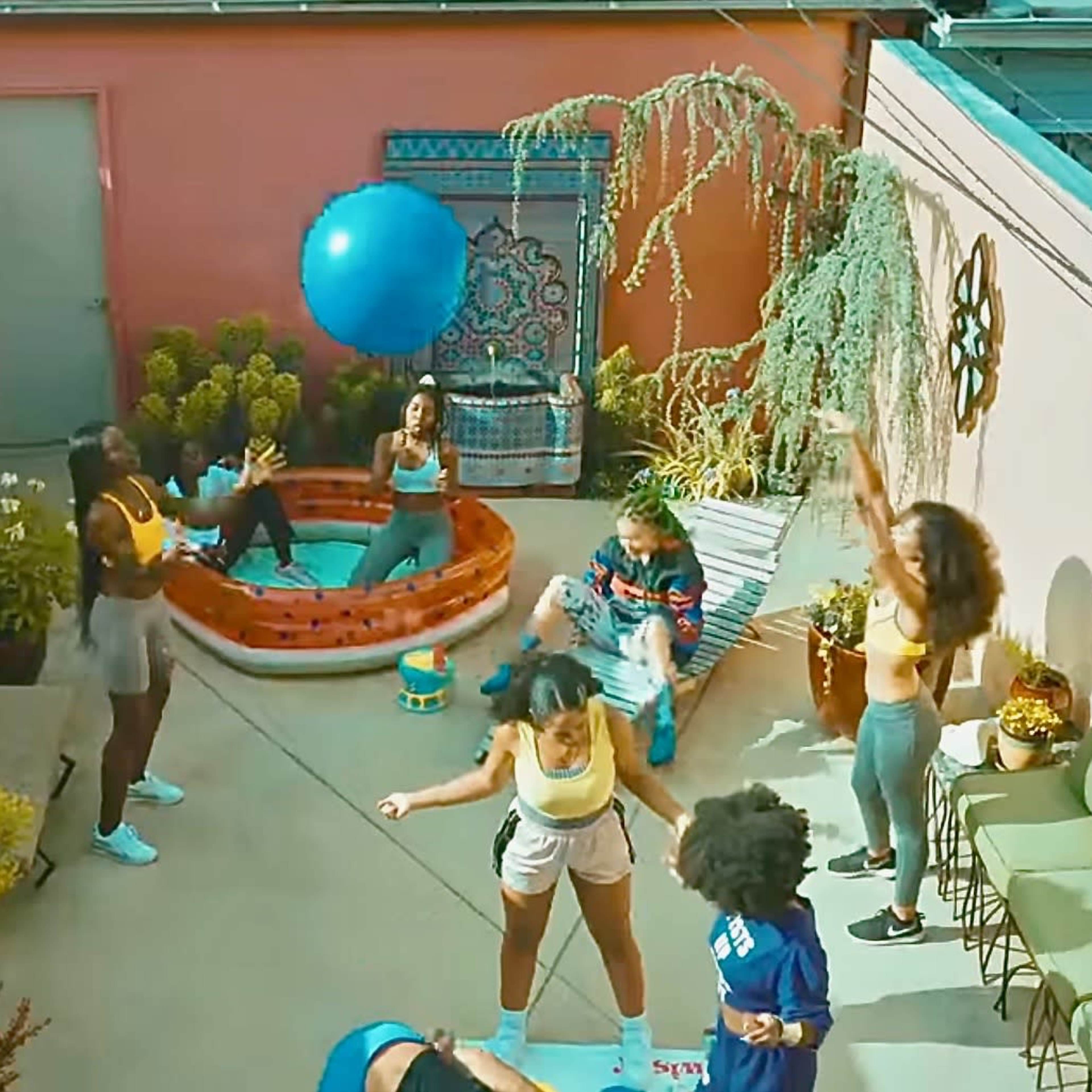 A group of people engages in playful activities in a brightly colored outdoor courtyard surrounded by plants and a small inflatable pool.