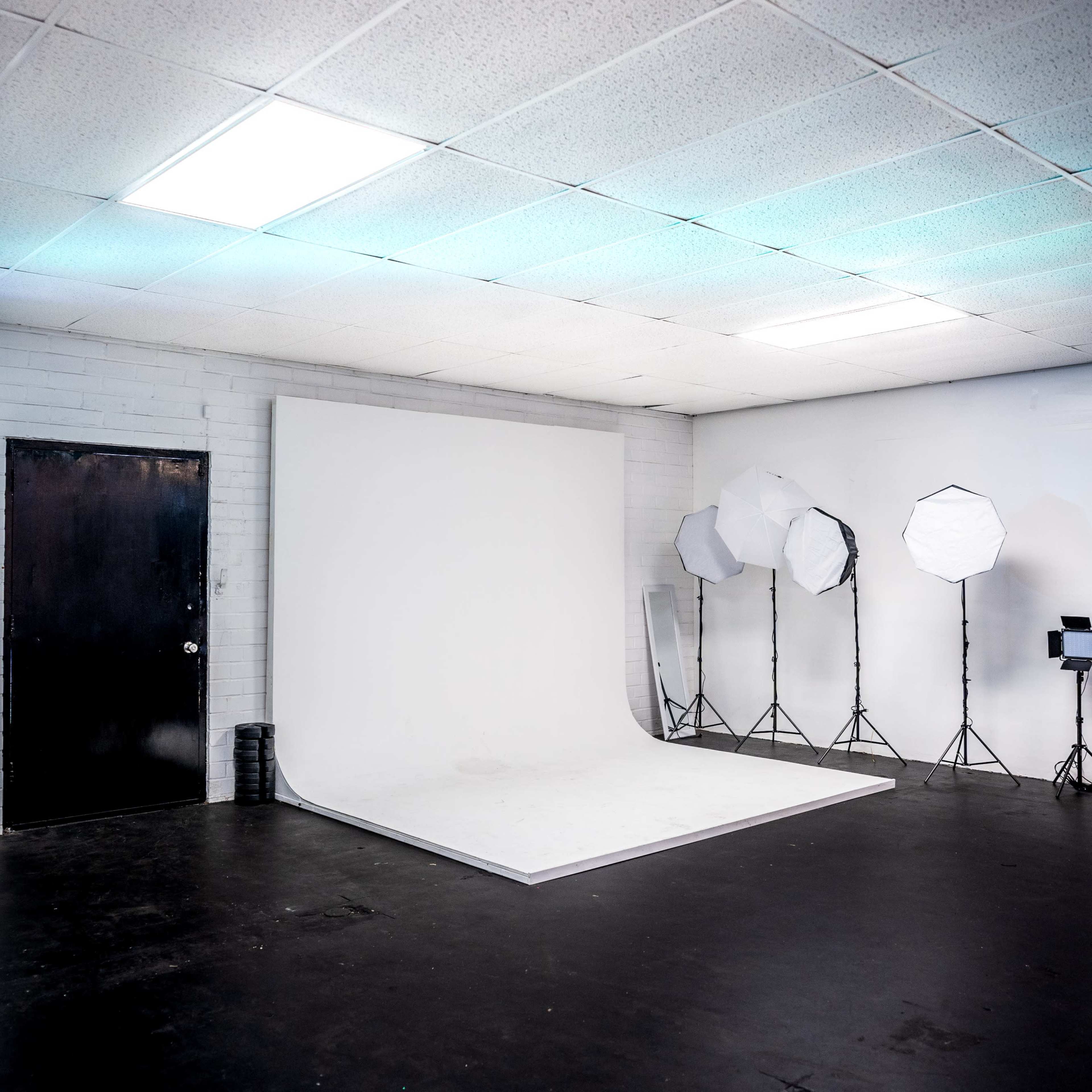 An empty photography studio with a white backdrop, black flooring, and lighting equipment set up on one side.
