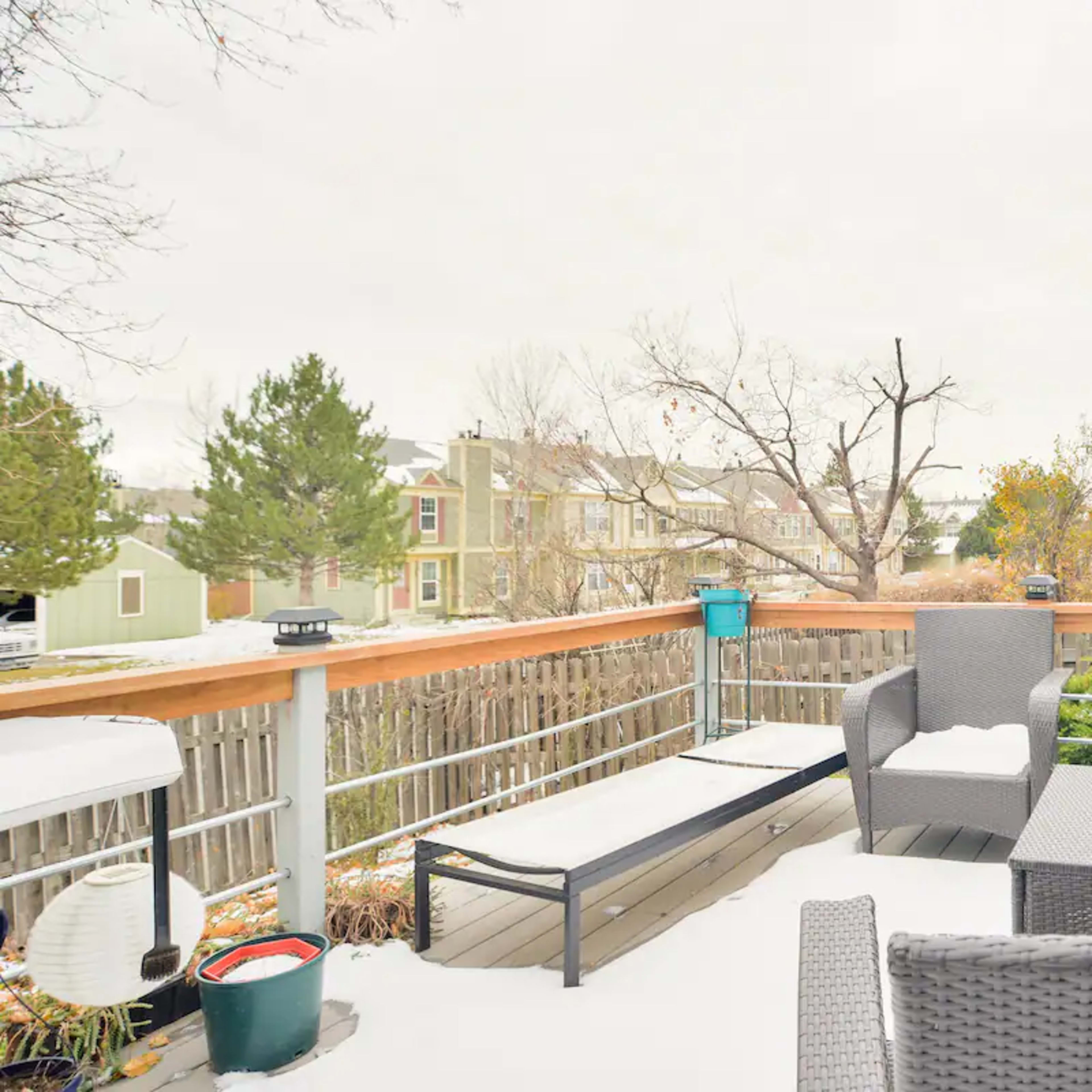 A wooden deck with outdoor furniture, including chairs and a lounge, surrounded by a suburban landscape with bare trees and nearby houses.