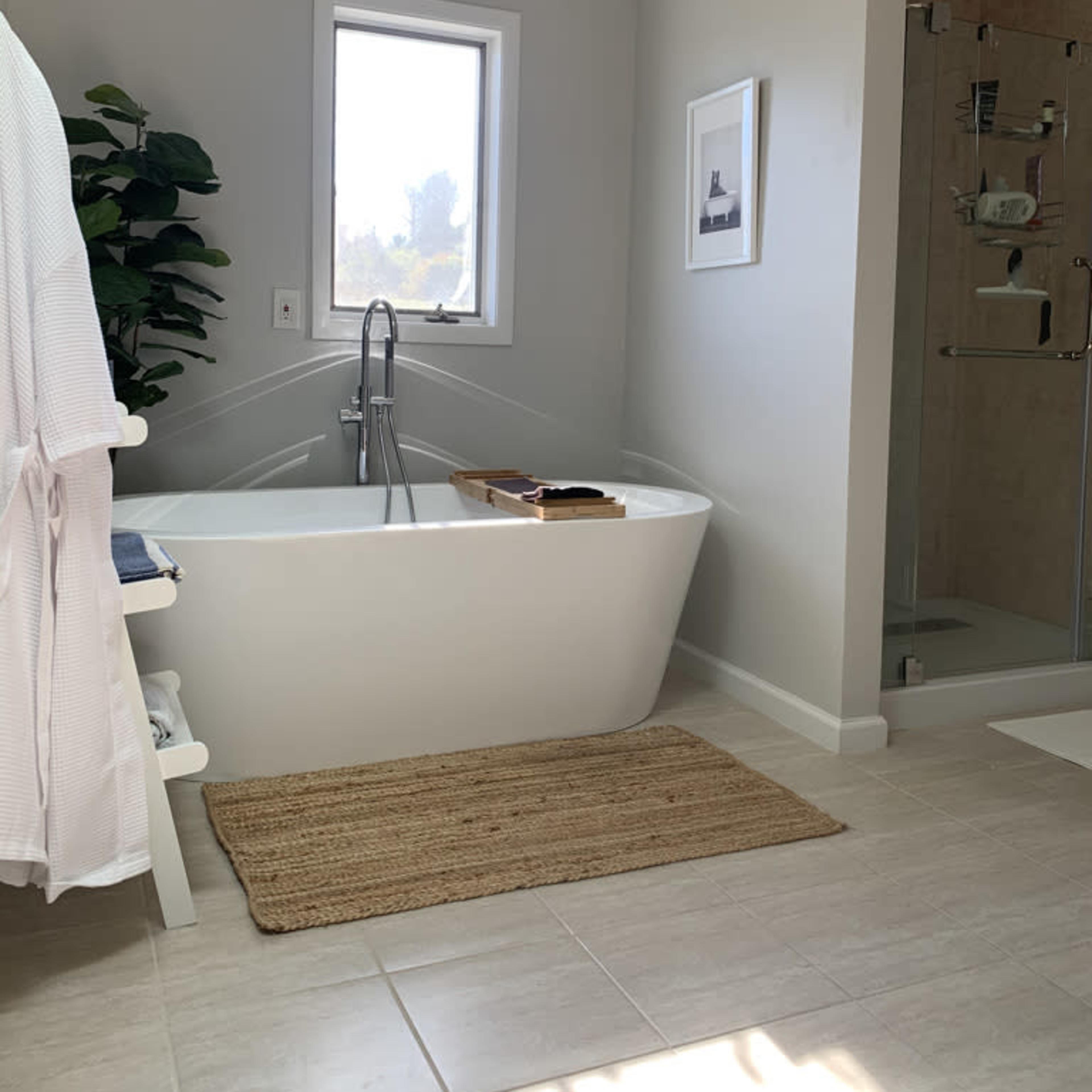 A modern bathroom featuring a freestanding bathtub, a glass shower enclosure, a window, and a woven rug on tiled flooring.