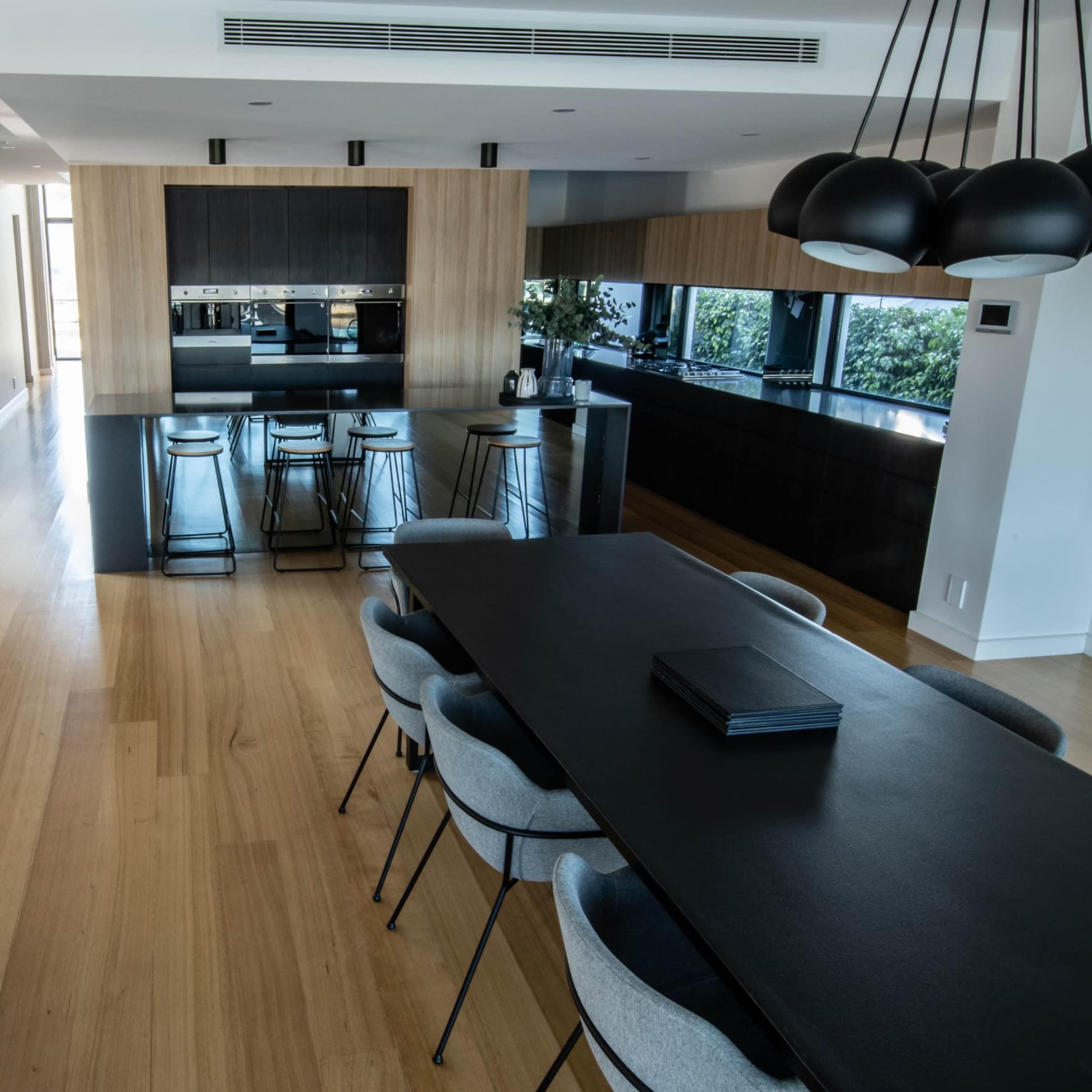 A modern open-concept kitchen and dining area with wooden flooring, a long dining table, and sleek black cabinetry.