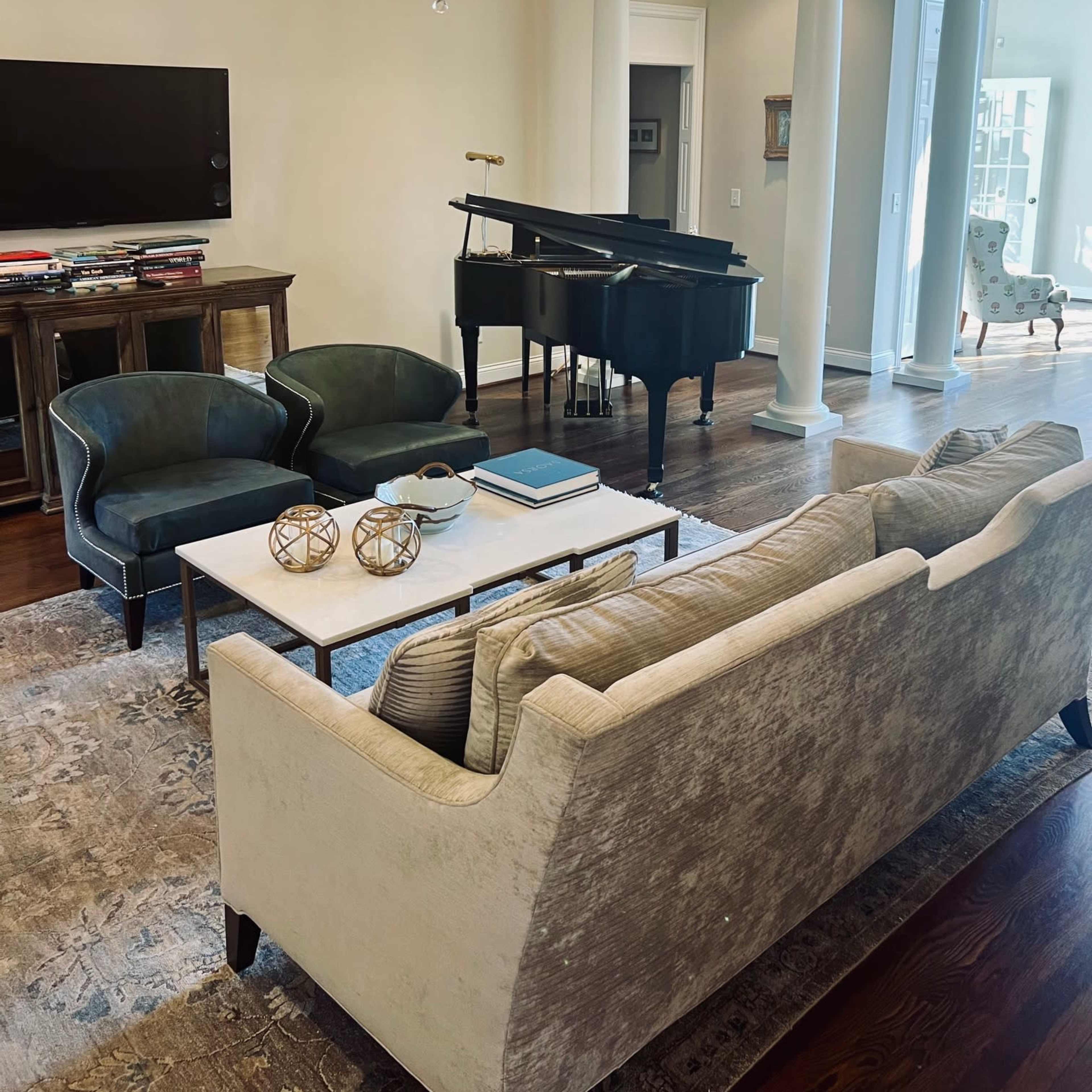 A well-furnished living room featuring a grand piano, a chandelier, a television mounted on the wall, and a coffee table with decorative objects.