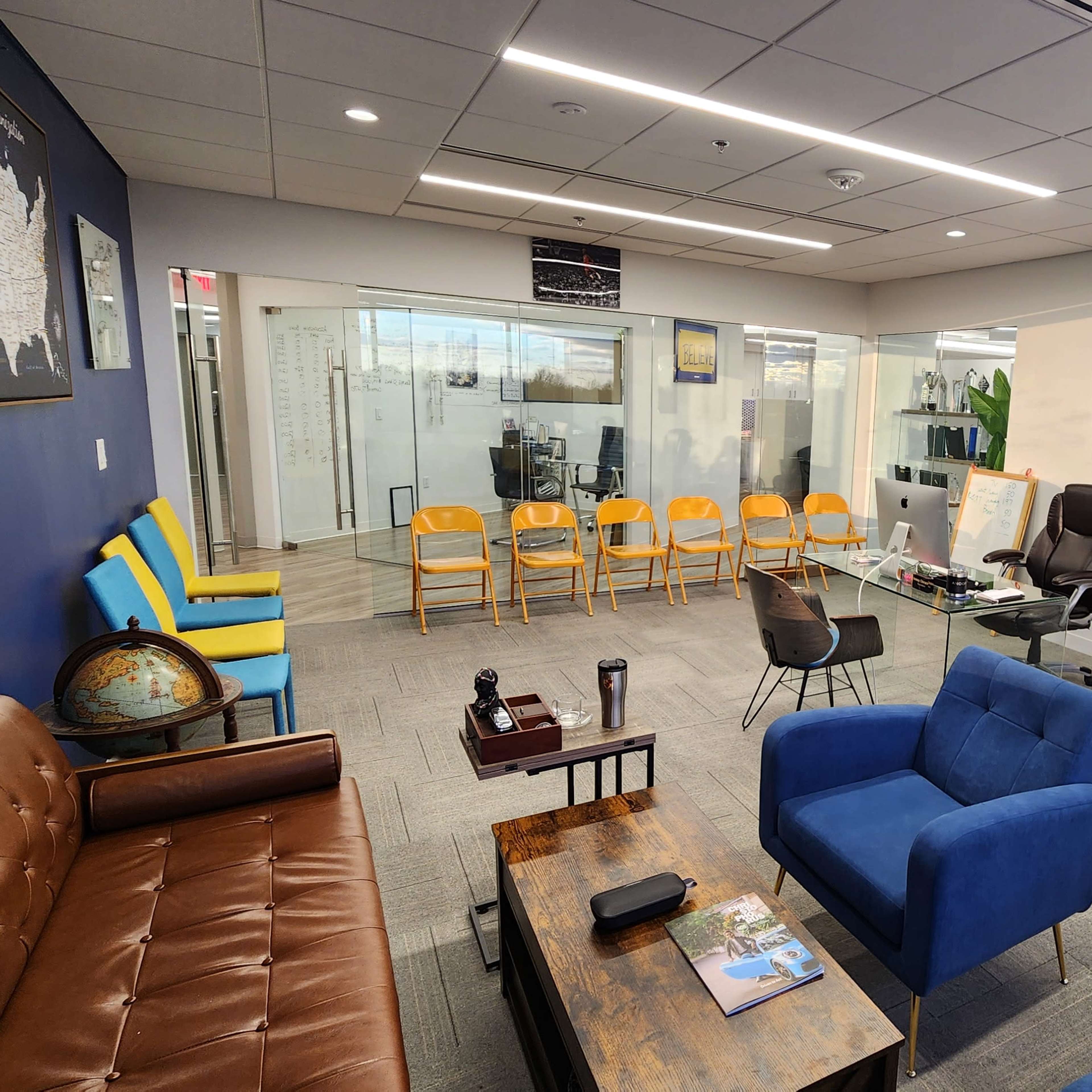 A modern office space with seating areas, a glass-walled meeting room, and colorful chairs arranged in rows.