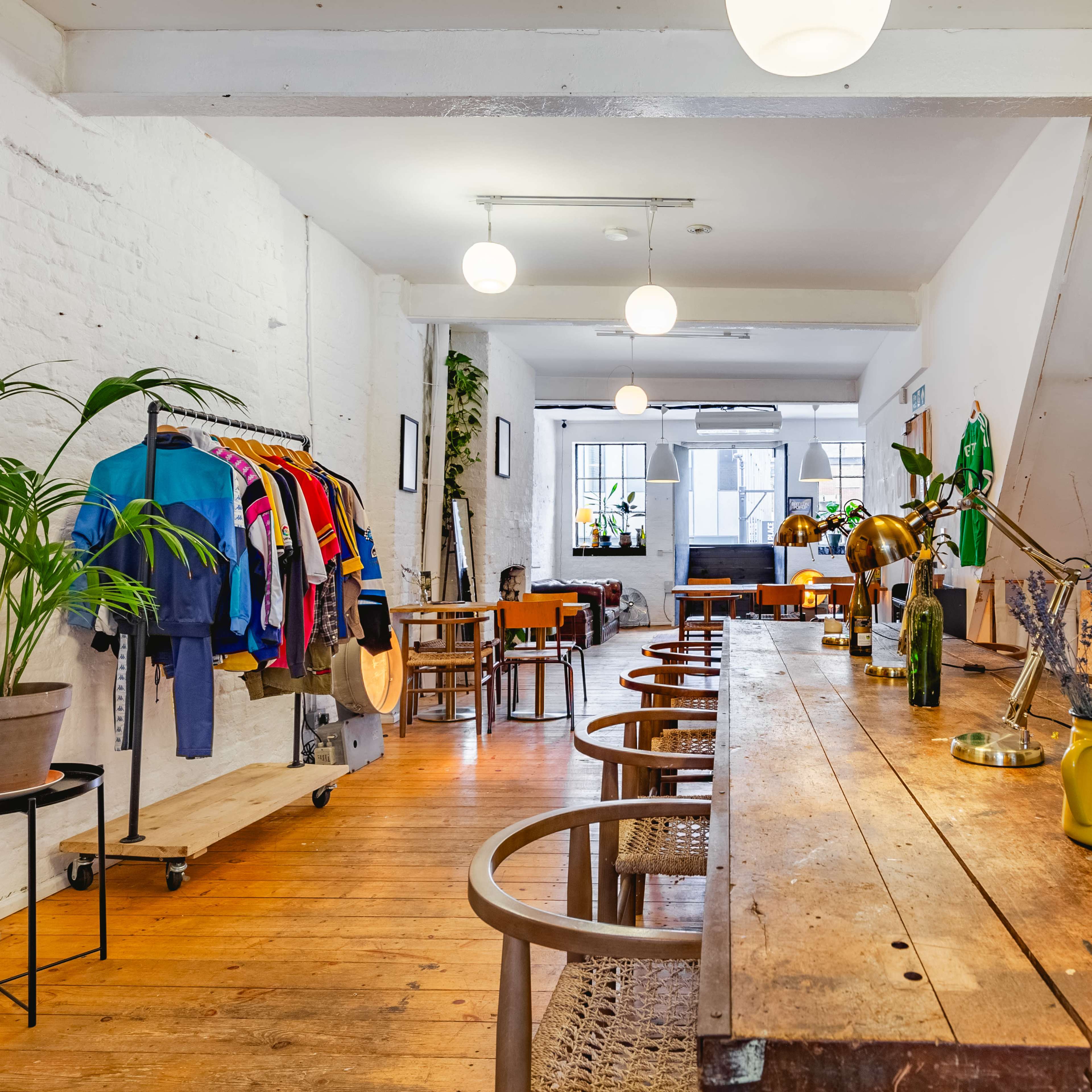 A spacious indoor area features a long wooden table, colorful clothing displayed on racks, and large windows providing ample light.