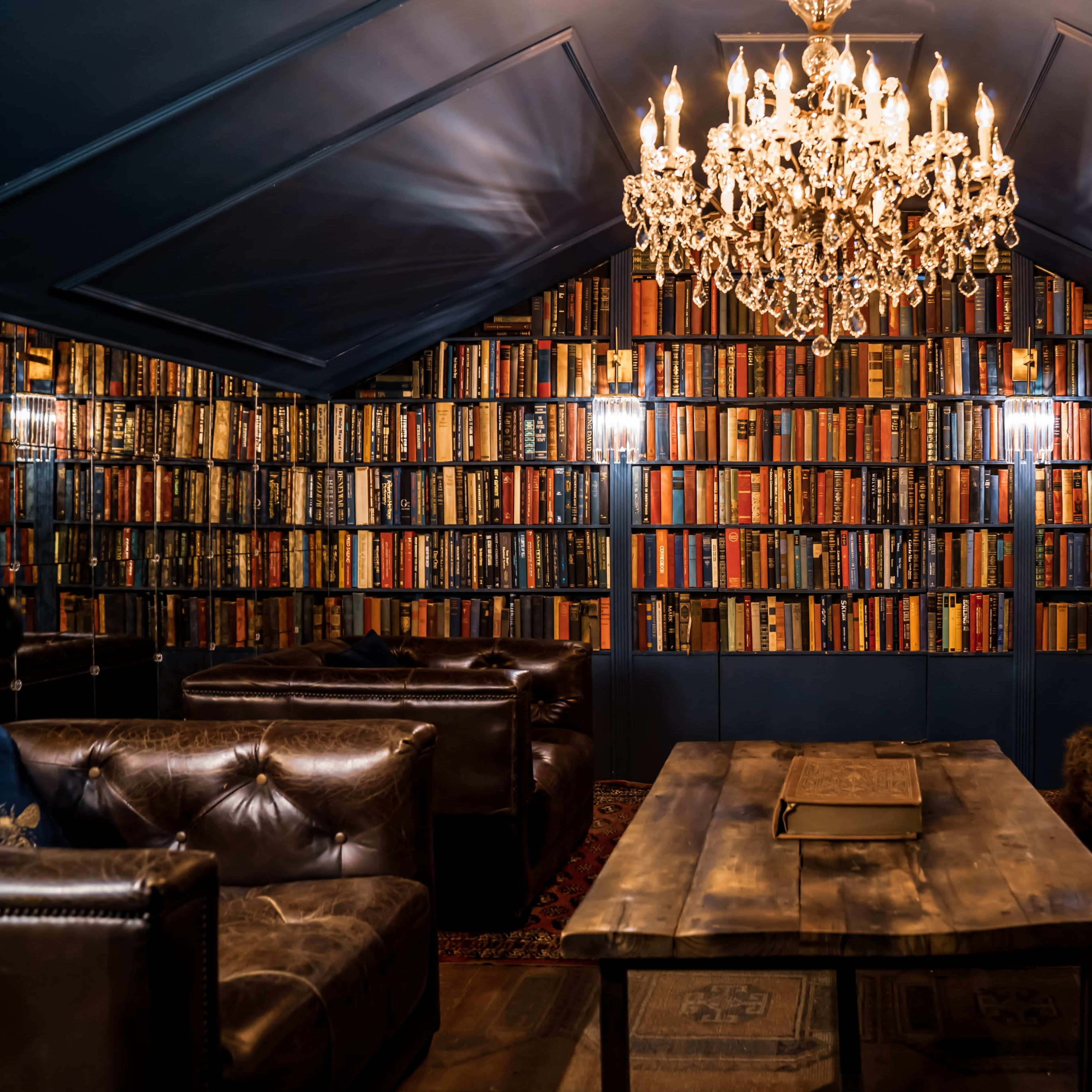 A cozy library features floor-to-ceiling shelves filled with colorful books, a wooden table in the center, and a crystal chandelier overhead.