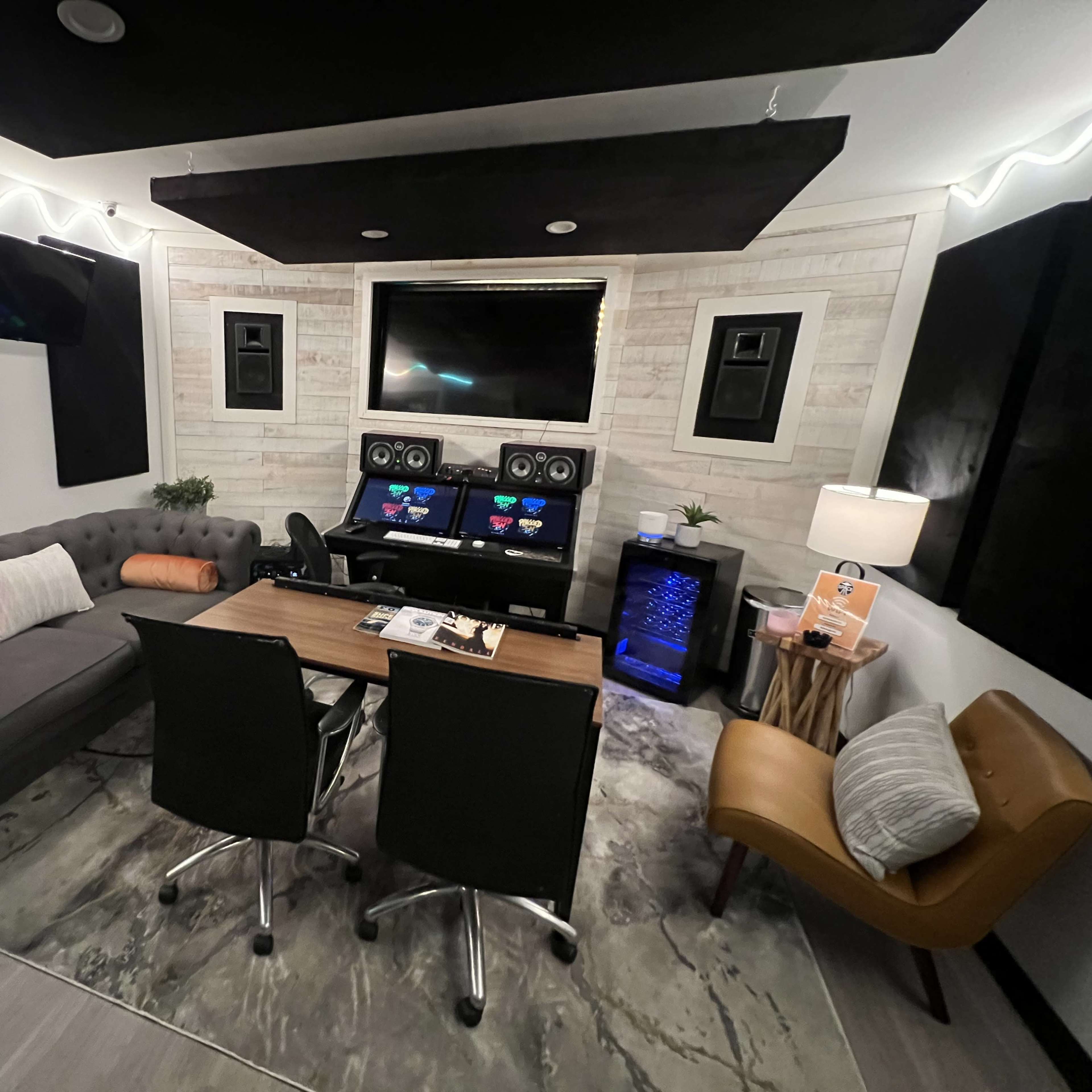A modern recording studio with a gray sofa, a wooden table with two black chairs, and sound equipment mounted on the walls, all set against a light-colored wood panel background.