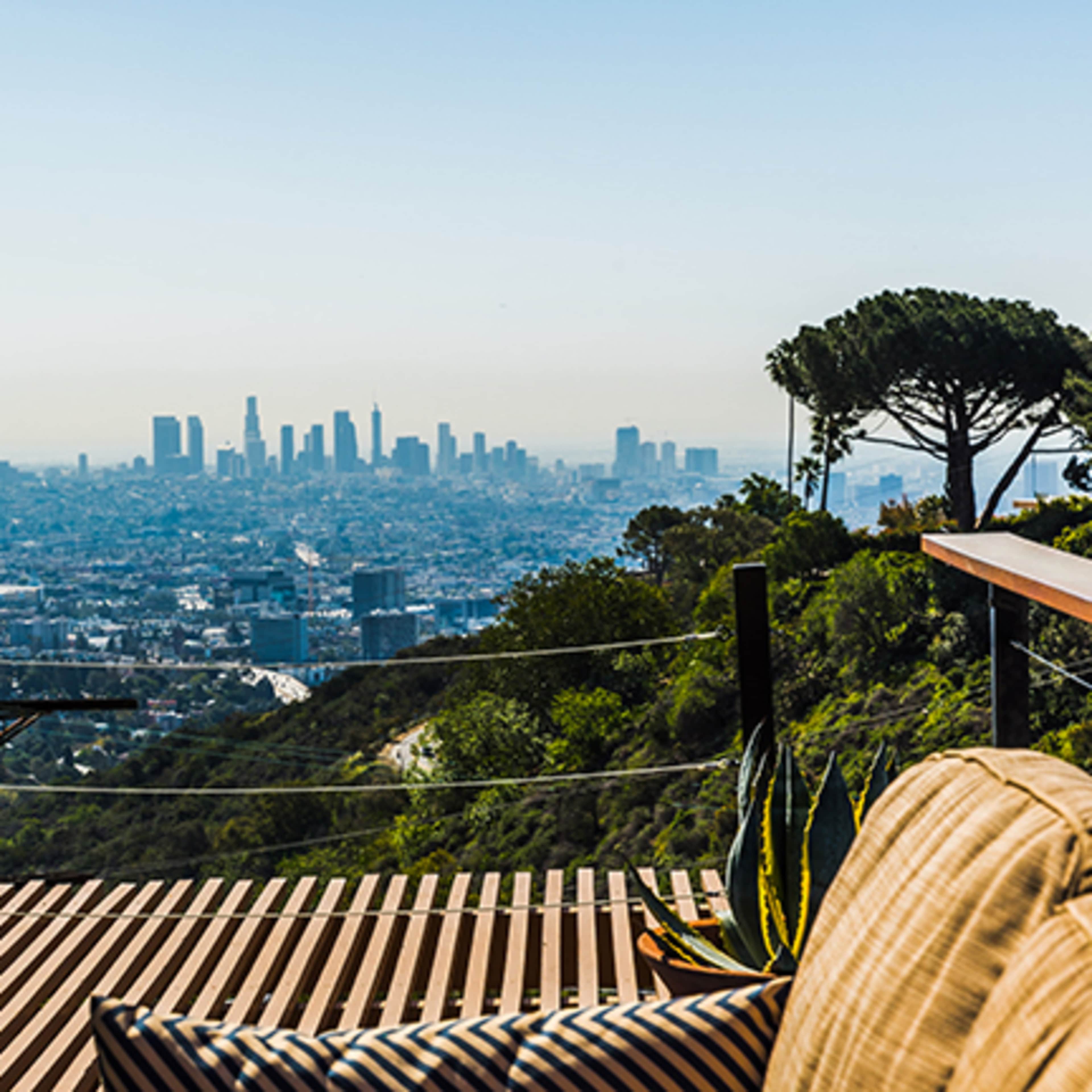 A view of the Los Angeles skyline set against a clear sky, with a foreground of outdoor seating and greenery.