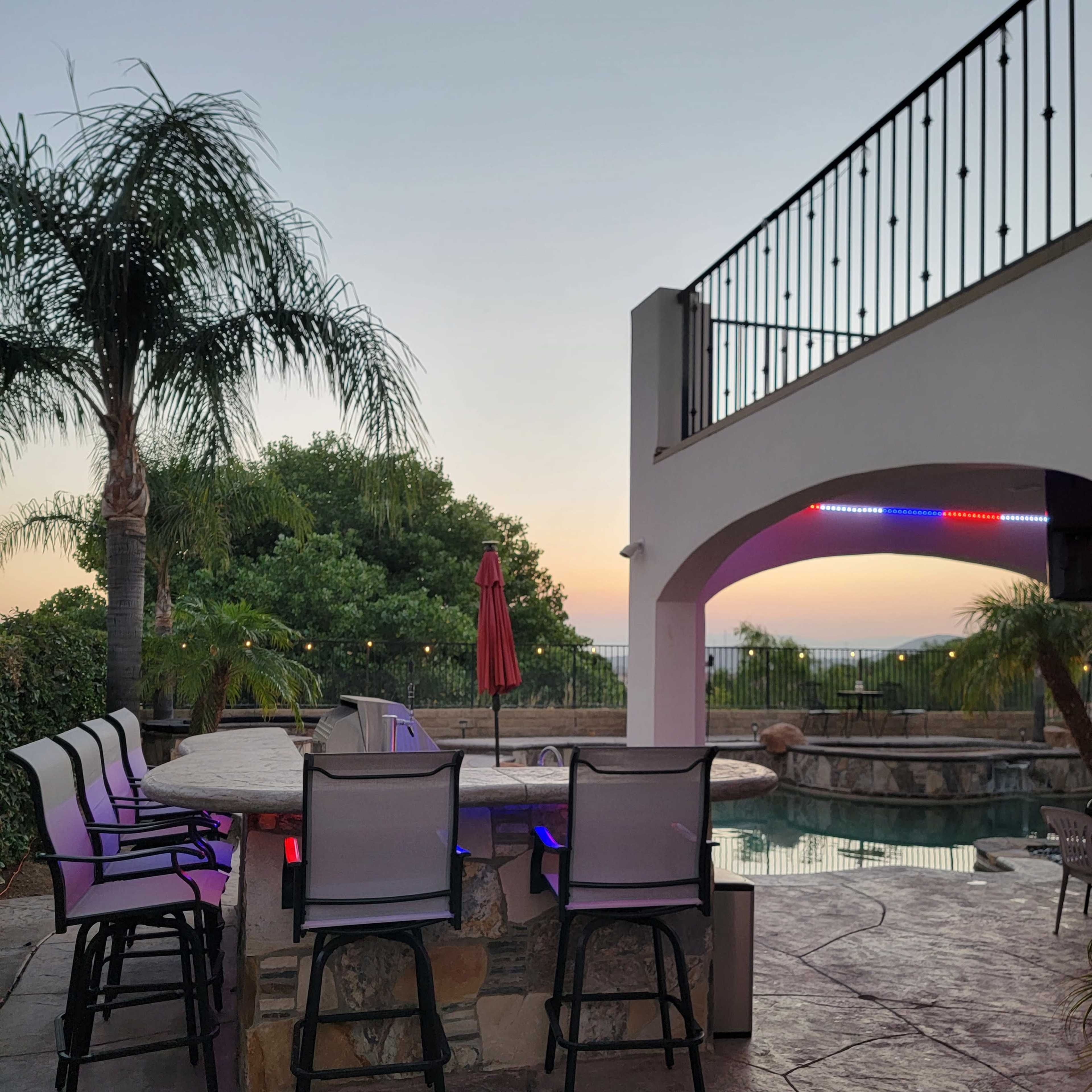 A backyard with bar stools surrounding a stone bar near a swimming pool, illuminated by lights, with palm trees and a sunset in the background.