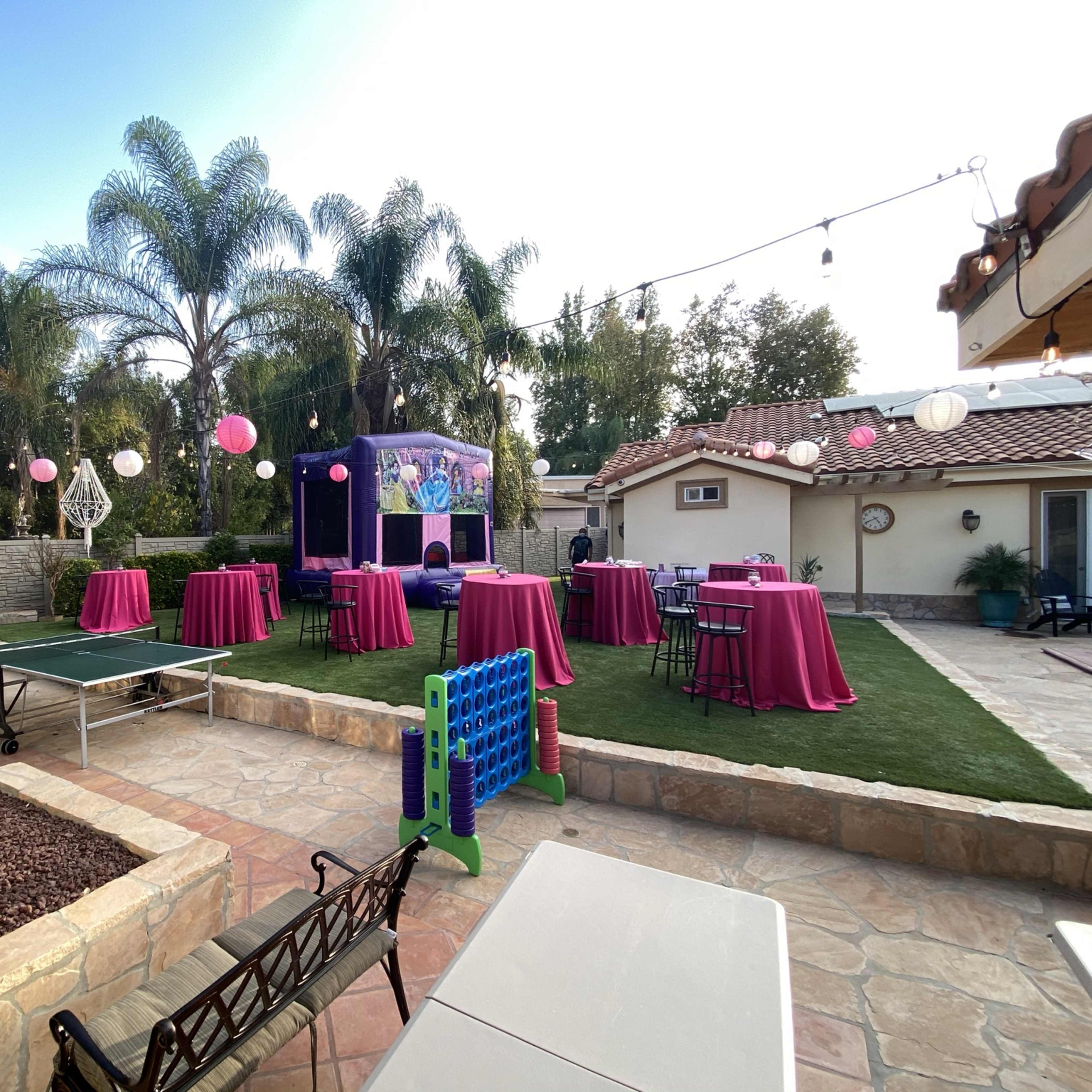 A backyard party setup features tables with pink tablecloths, string lights, and decorative elements surrounded by palm trees.