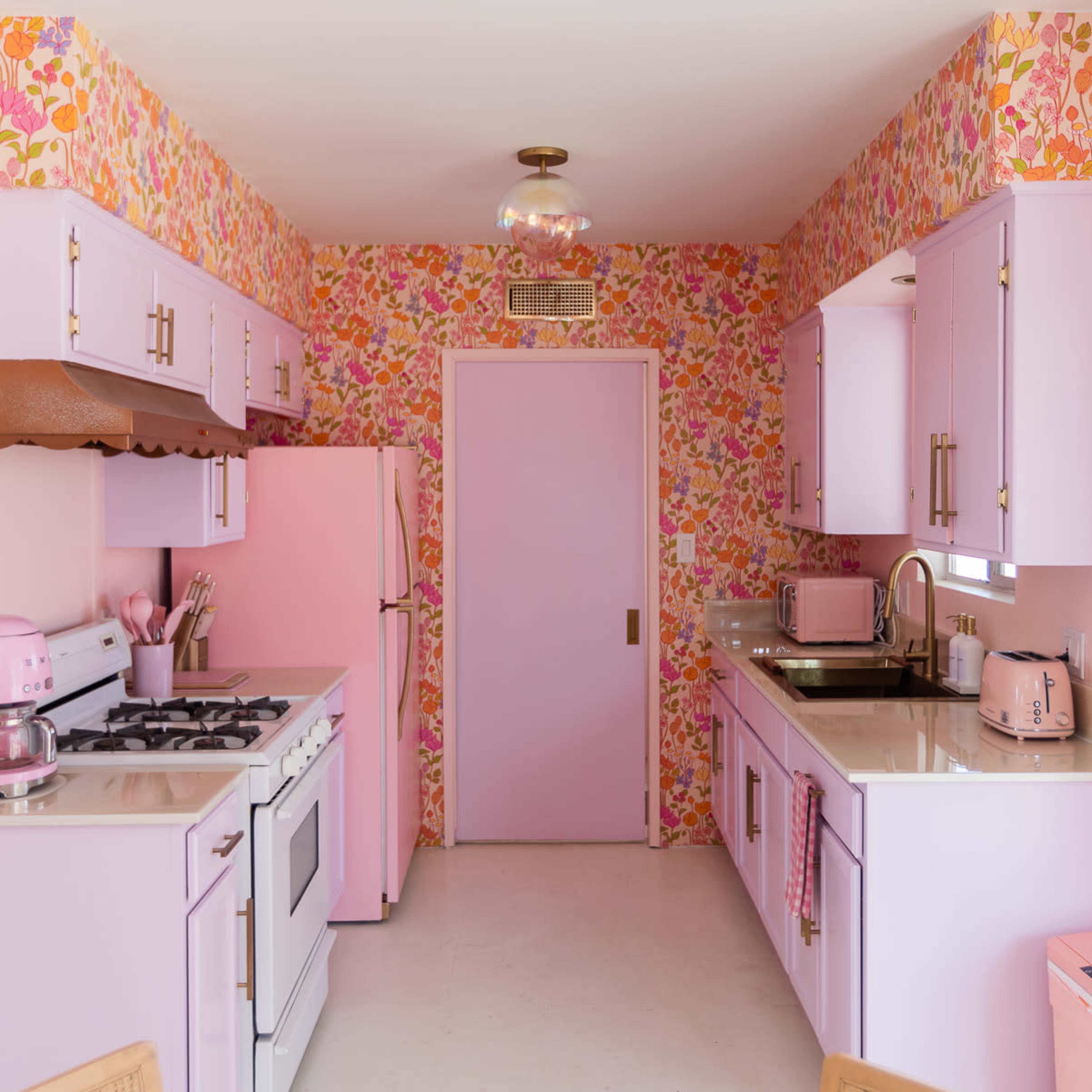 The kitchen features pastel pink and floral wallpaper, with lavender cabinets and appliances, including a pink refrigerator and toaster.