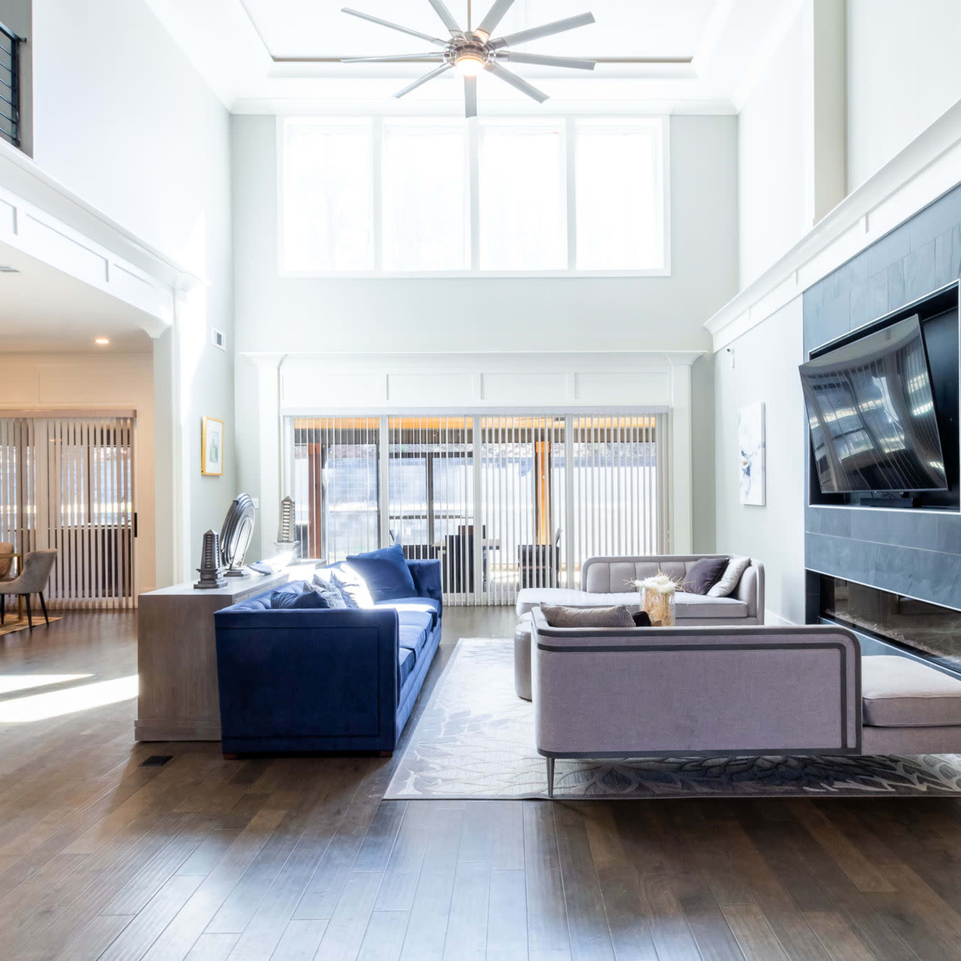 The spacious living area features a blue sofa and a gray sectional, with large windows allowing natural light to fill the room.