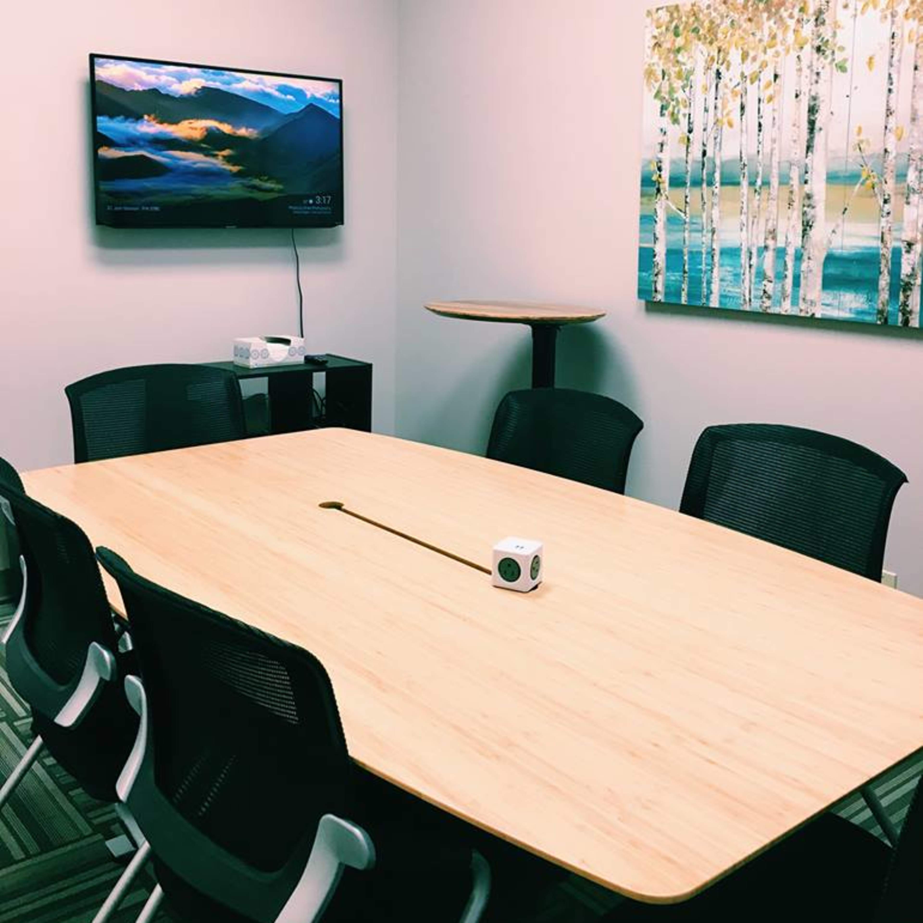 A conference room features a large wooden table surrounded by chairs, a wall-mounted television displaying a landscape, and a piece of artwork depicting a forest scene.