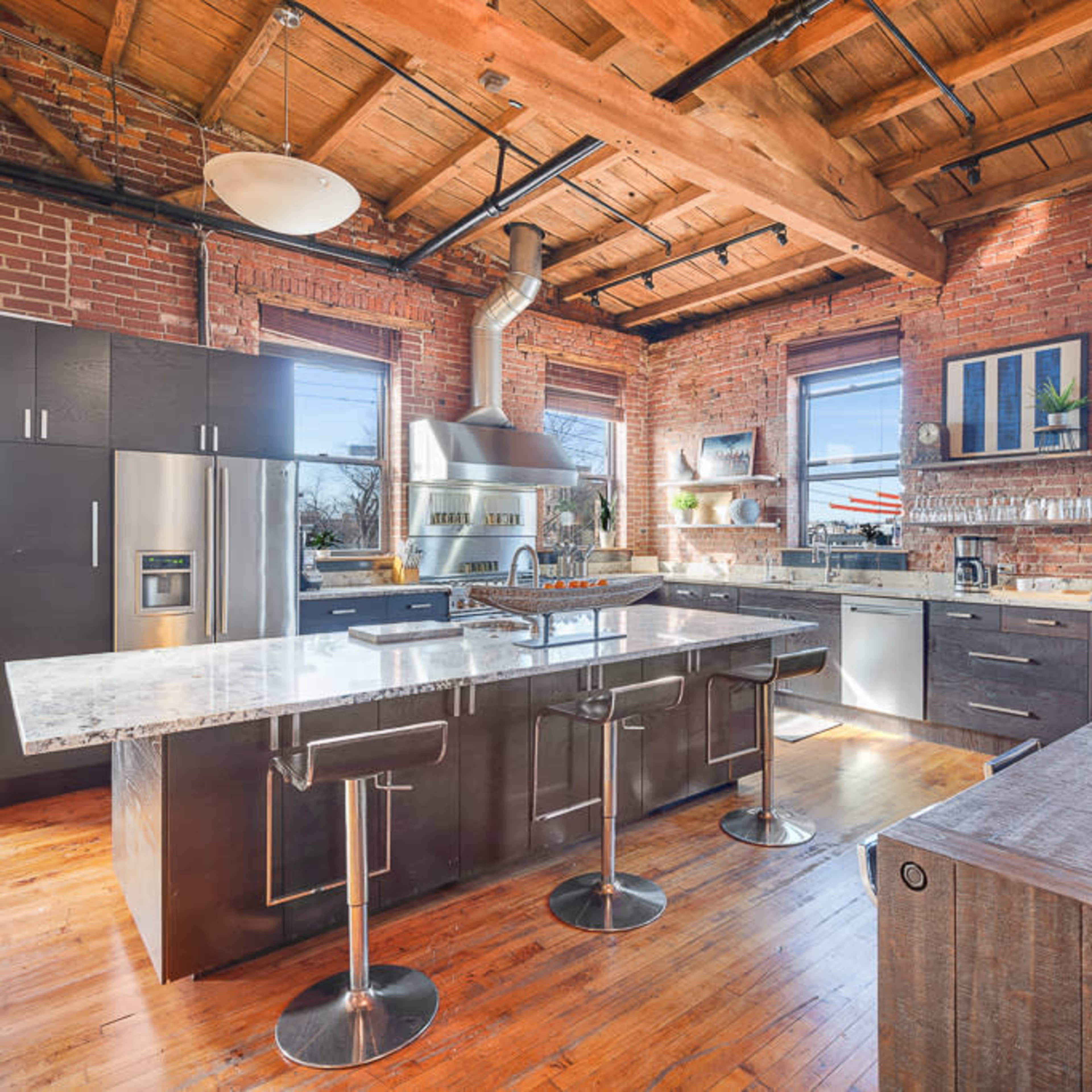 A modern kitchen with exposed brick walls, wooden beams, and a large island featuring bar stools.