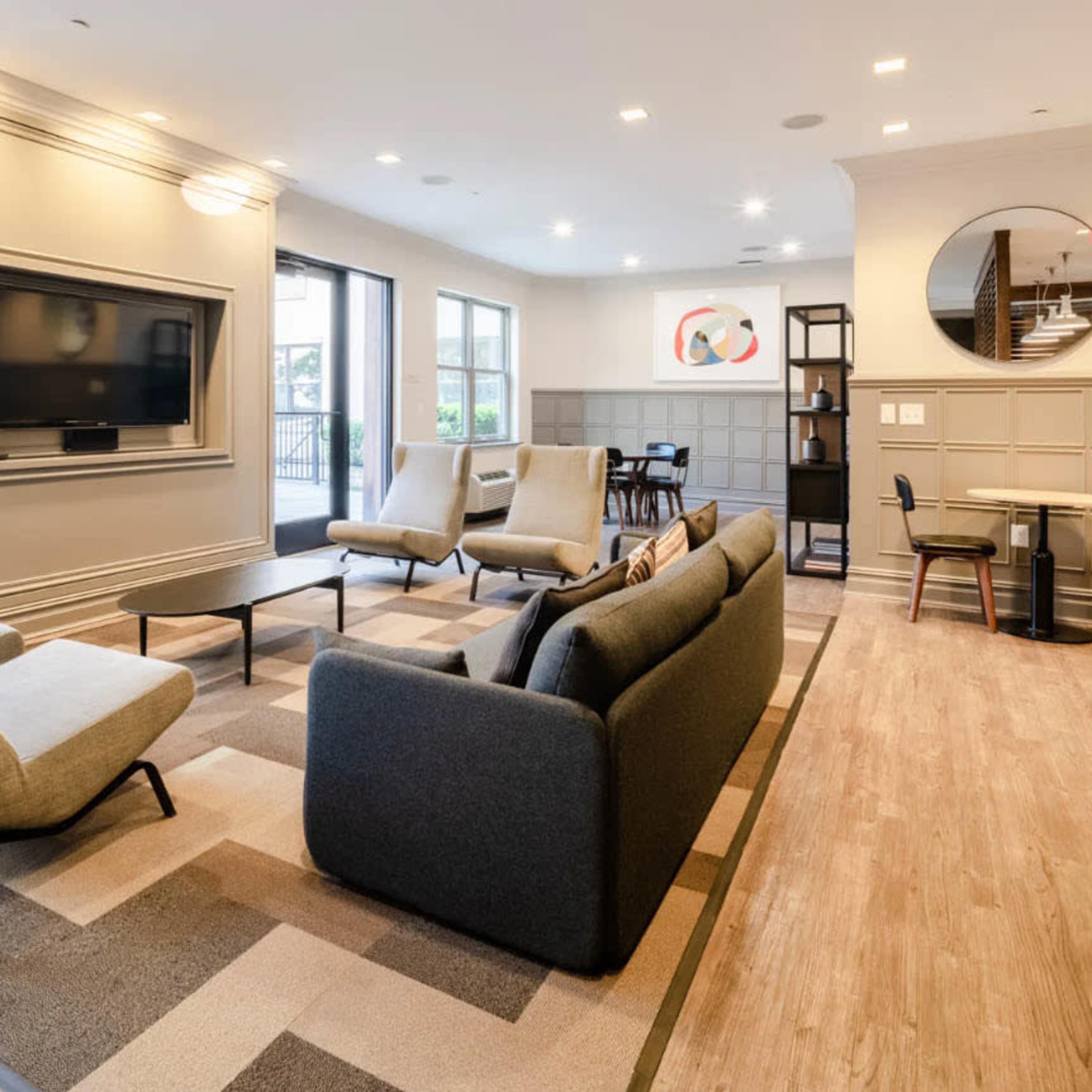 A modern lounge area with seating, a television mounted on the wall, and a small dining space in the background.