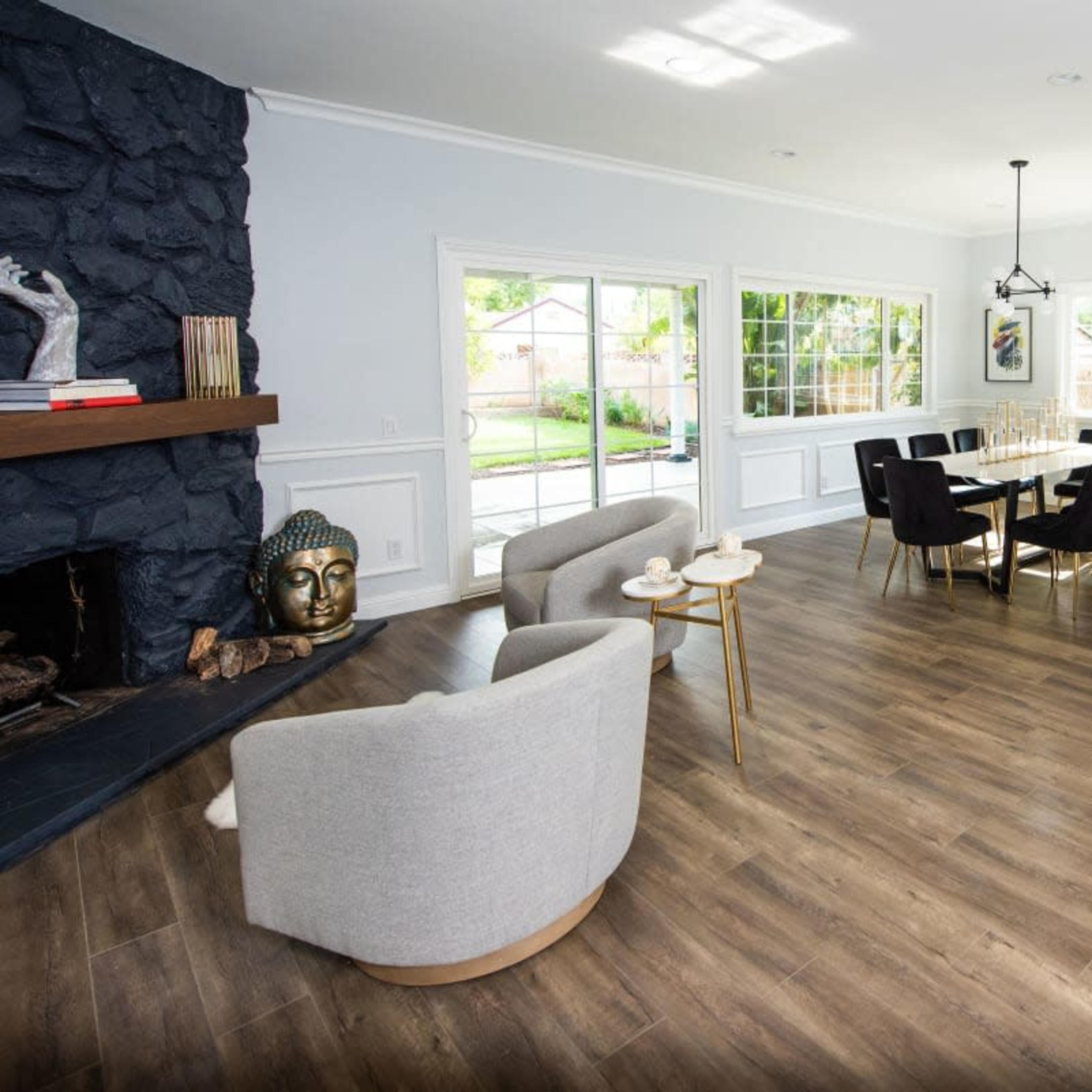 A spacious living area features a dark stone fireplace, a dining table with chairs, and a blue sofa accented with a throw pillow, all illuminated by natural light from large windows.