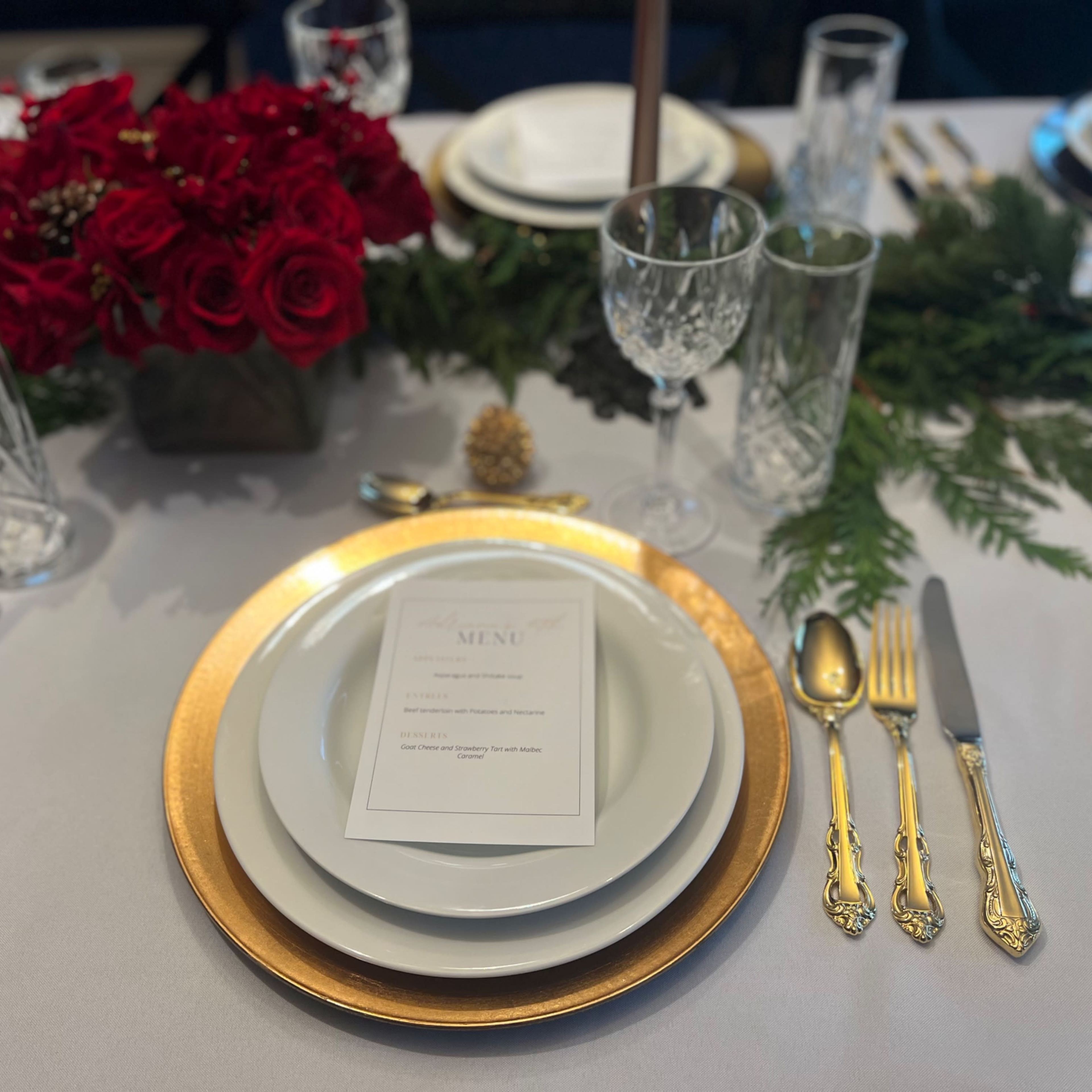 A beautifully arranged dining table features a gold-rimmed plate, a menu card, crystal glassware, silver utensils, and a centerpiece of red roses surrounded by greenery.