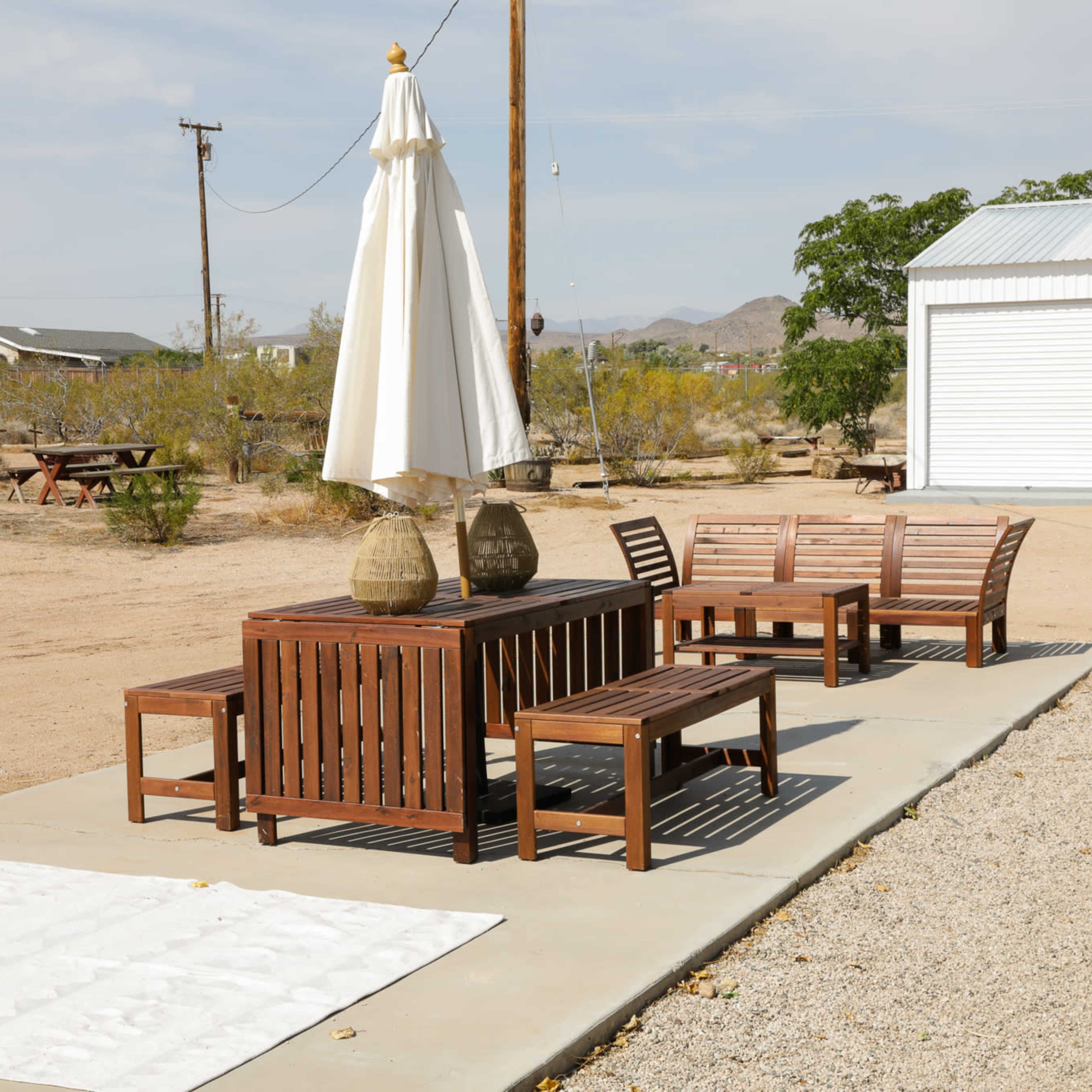 A wooden patio set with an umbrella is positioned on a concrete surface in a desert landscape near a storage building.