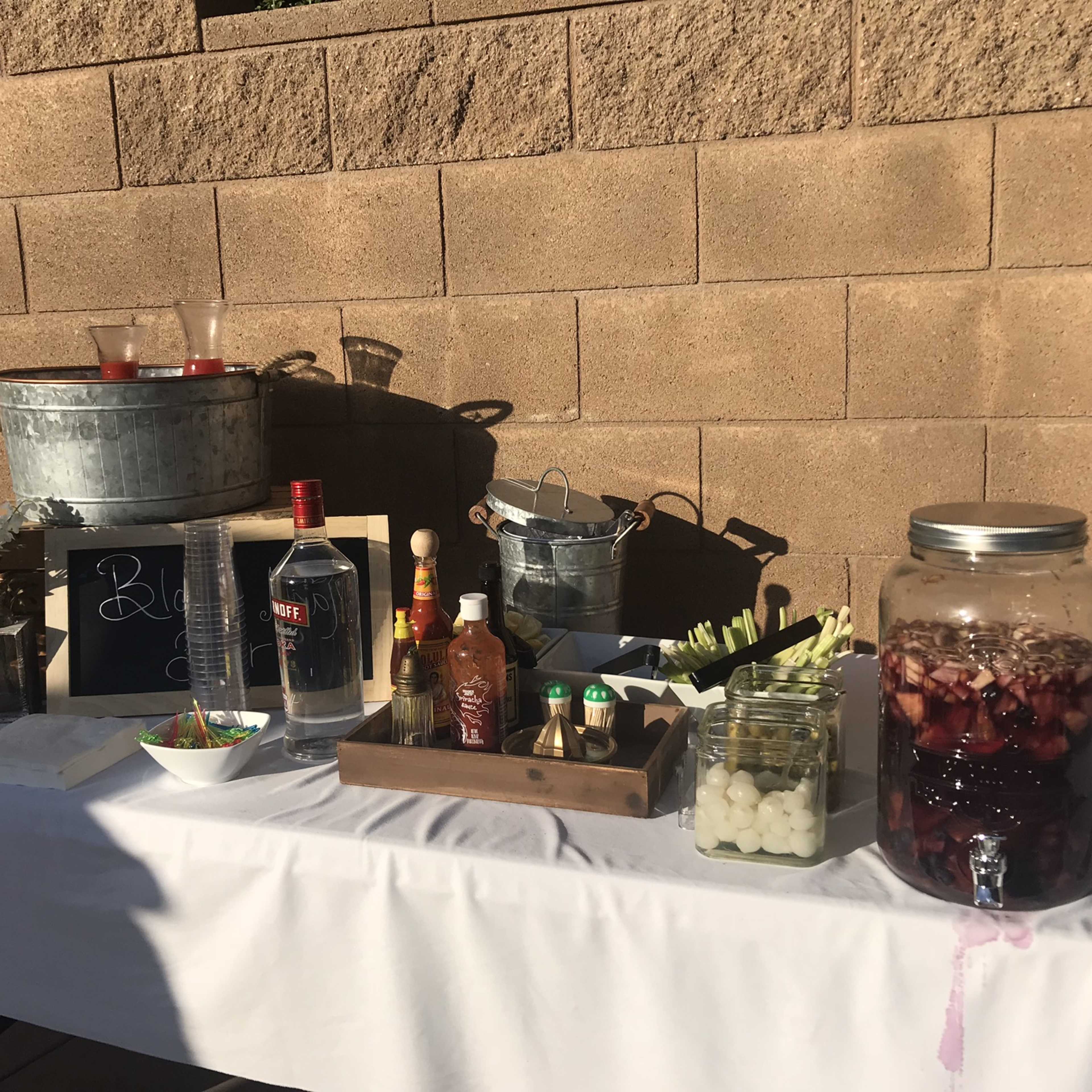 A table set up with various bottles of alcohol, mixers, and garnishes, with a chalkboard sign displaying the word "Beverages" against a textured wall.