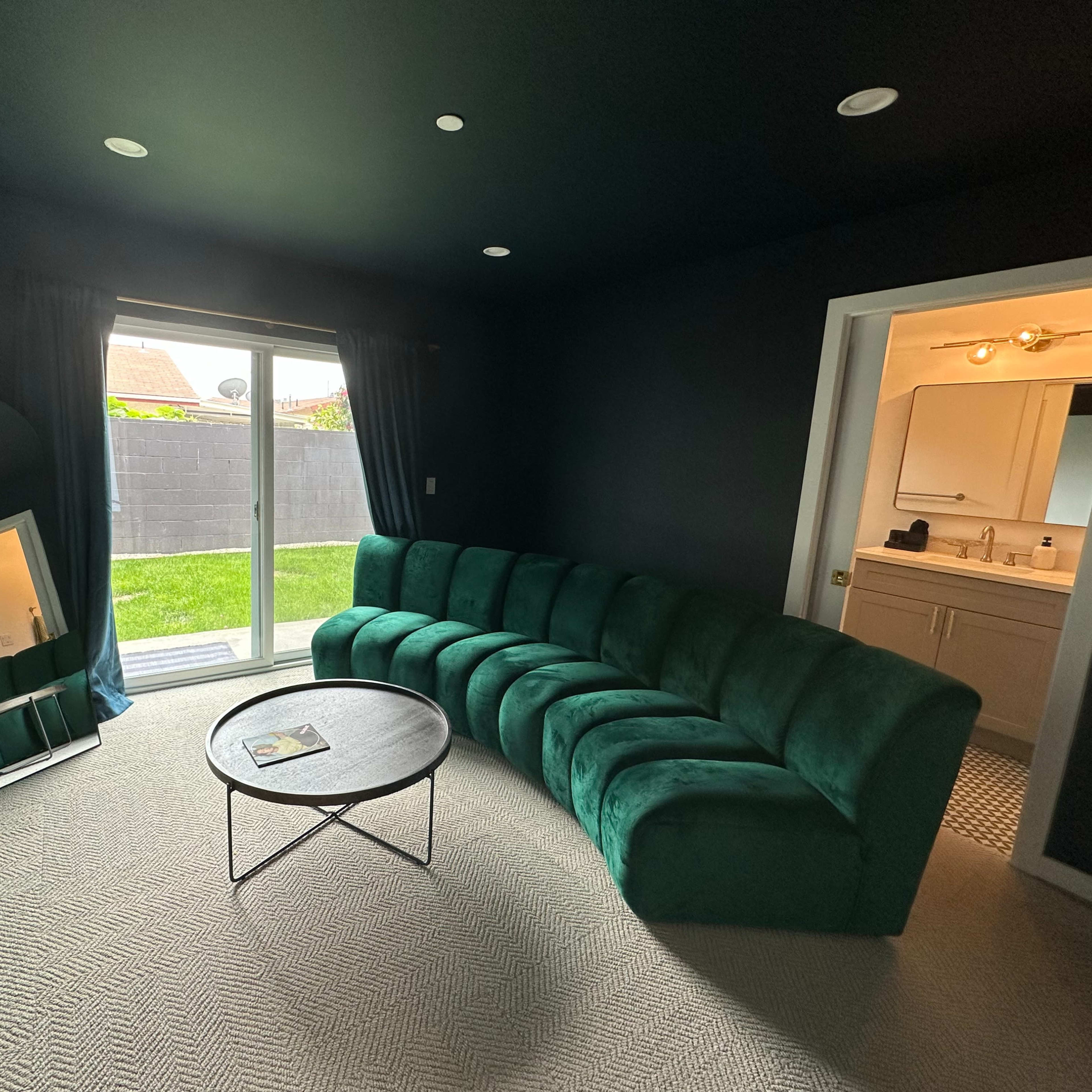 A darkly painted room with a curved green sofa, a round table, a large mirror, and a door leading to a well-lit area with a sliding glass door opening to a grassy outdoor space.