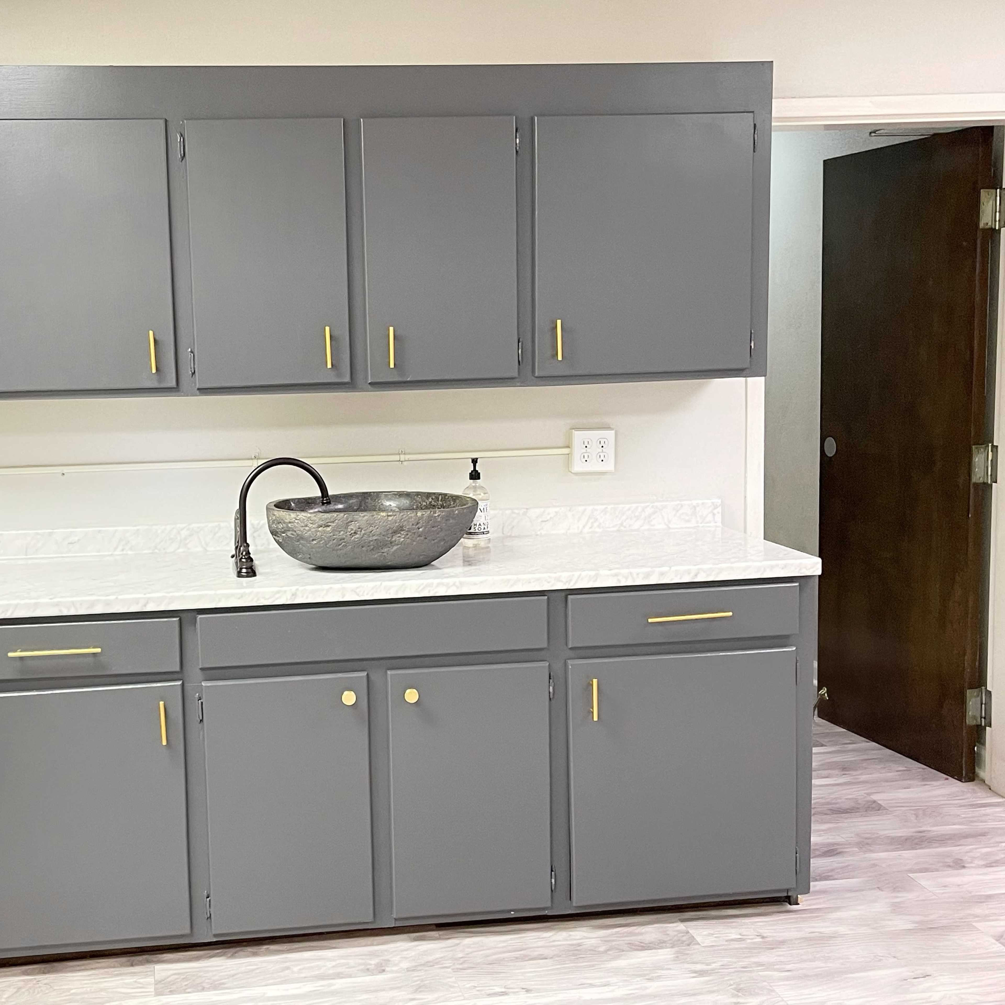 A gray kitchen cabinet set with a marble countertop features a round sink and a nearby white bar stool.