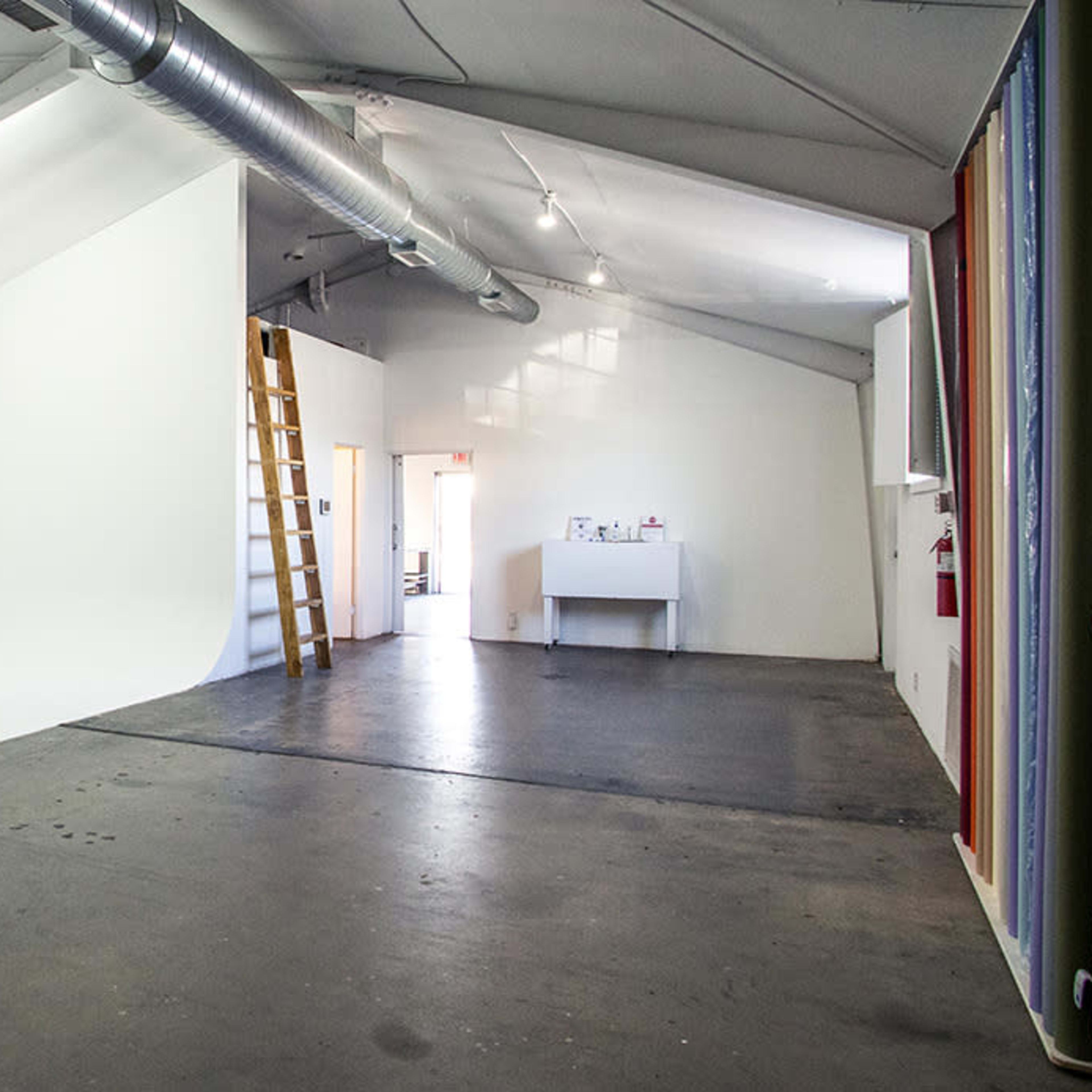 A spacious studio with a white backdrop on one side, a ladder leaning against the wall, and a small workstation in the background.