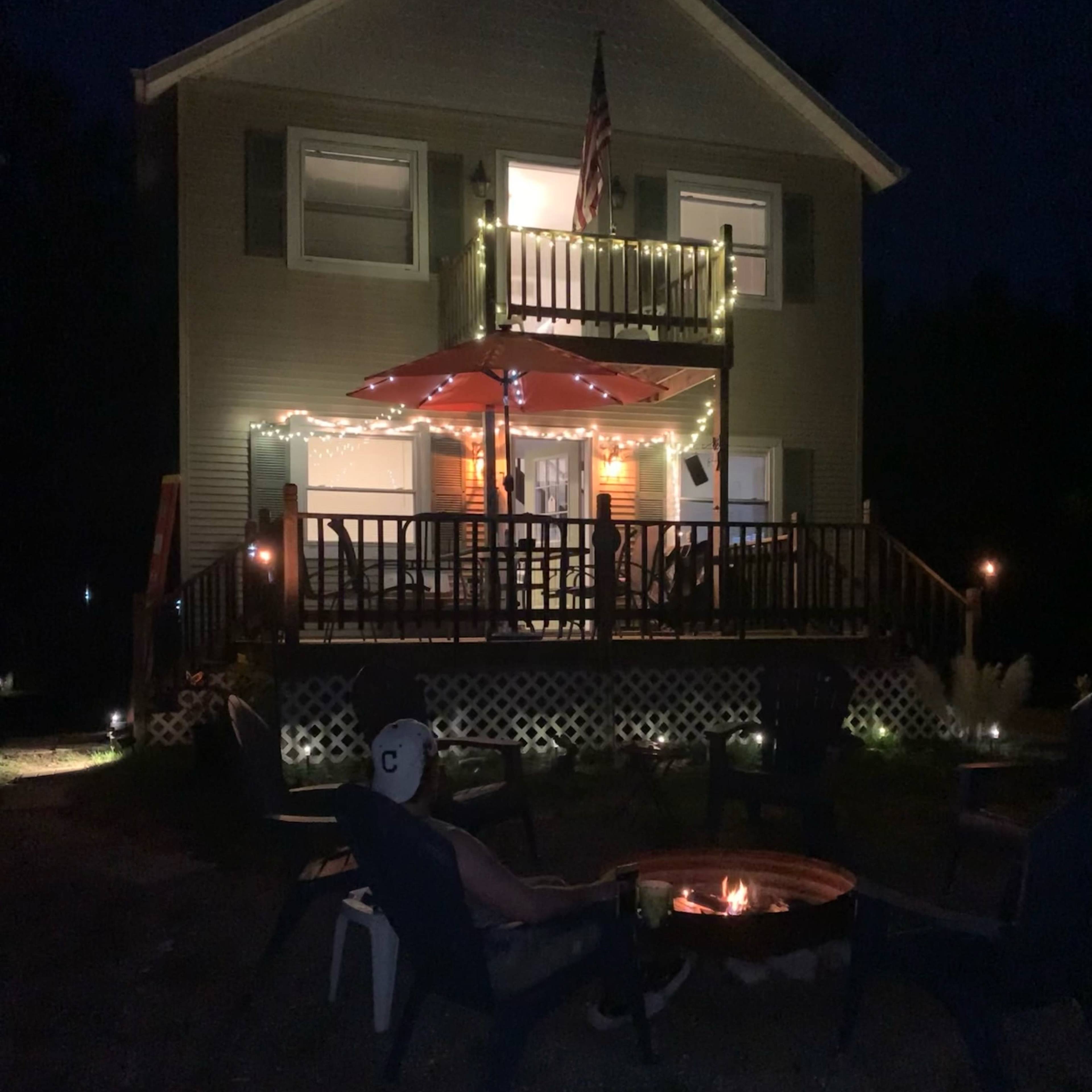 A person sits in front of a fire pit with a house illuminated by string lights and a large umbrella on the porch in the background.