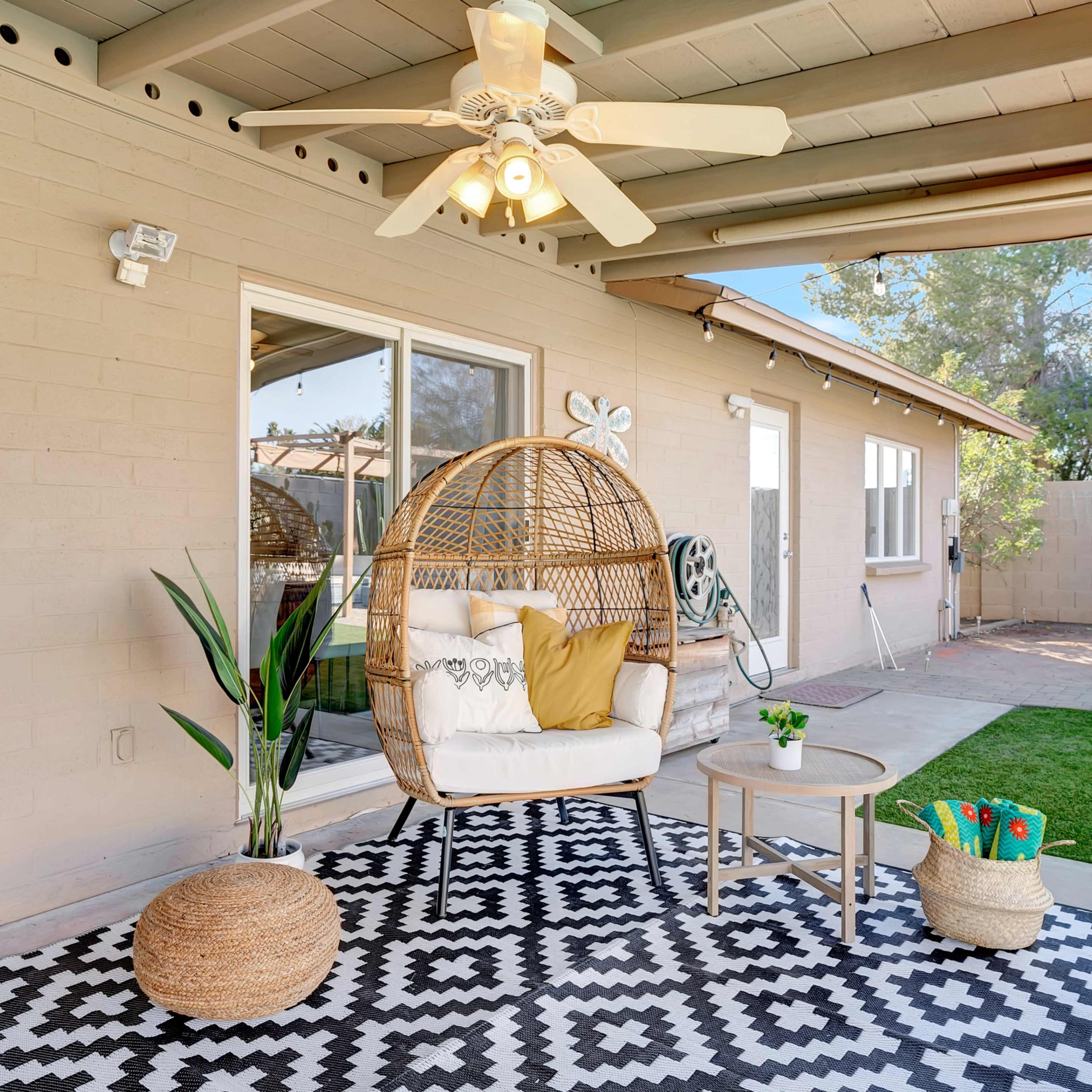 A covered patio features a hanging rattan chair with cushions, a small table, and potted plants, set on a geometric-patterned rug.
