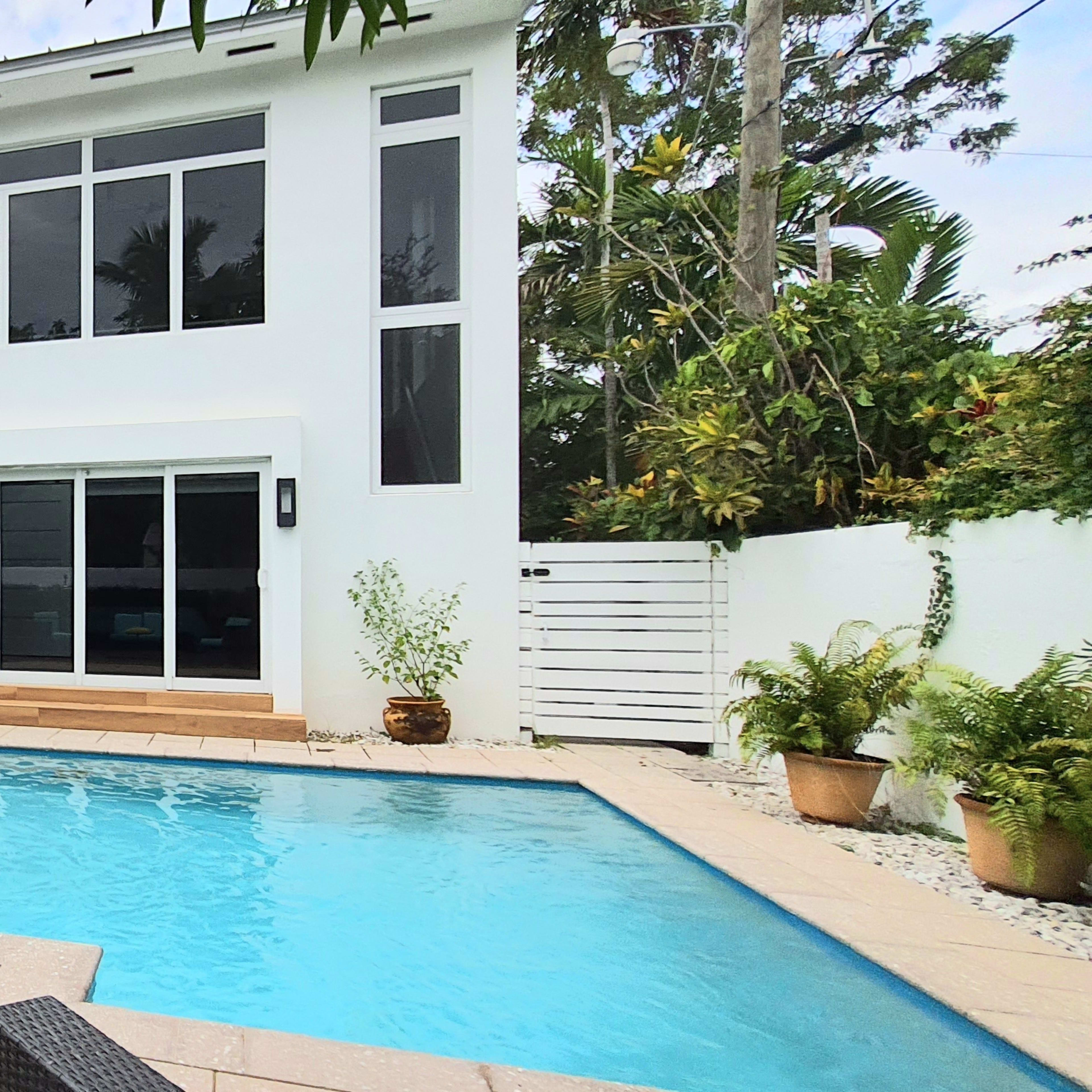 A modern home with a swimming pool, outdoor seating area, and large windows surrounded by tropical plants.