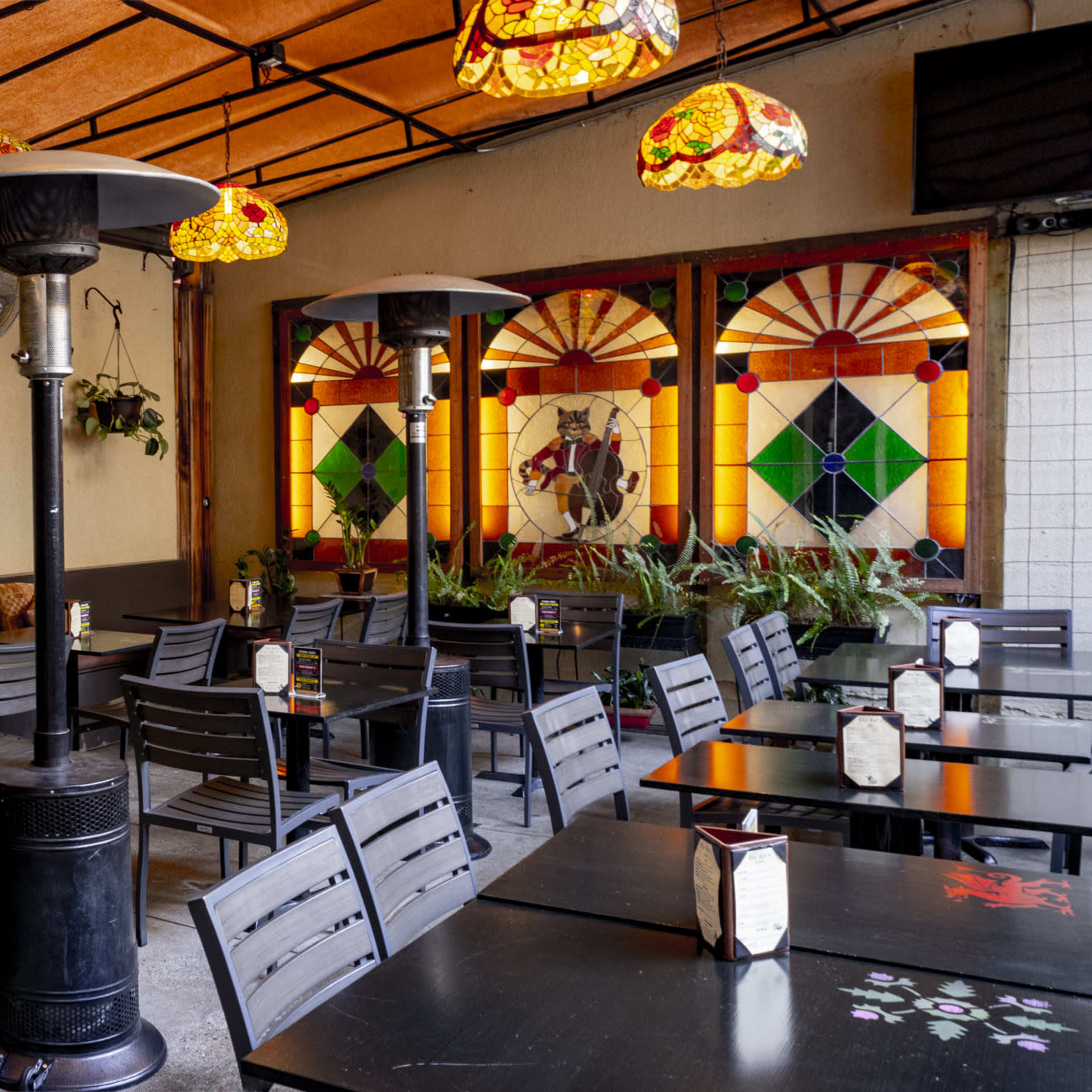 The interior of a restaurant features rows of black tables and chairs, decorative stained glass windows, and pendant lamps hanging from a wooden ceiling.