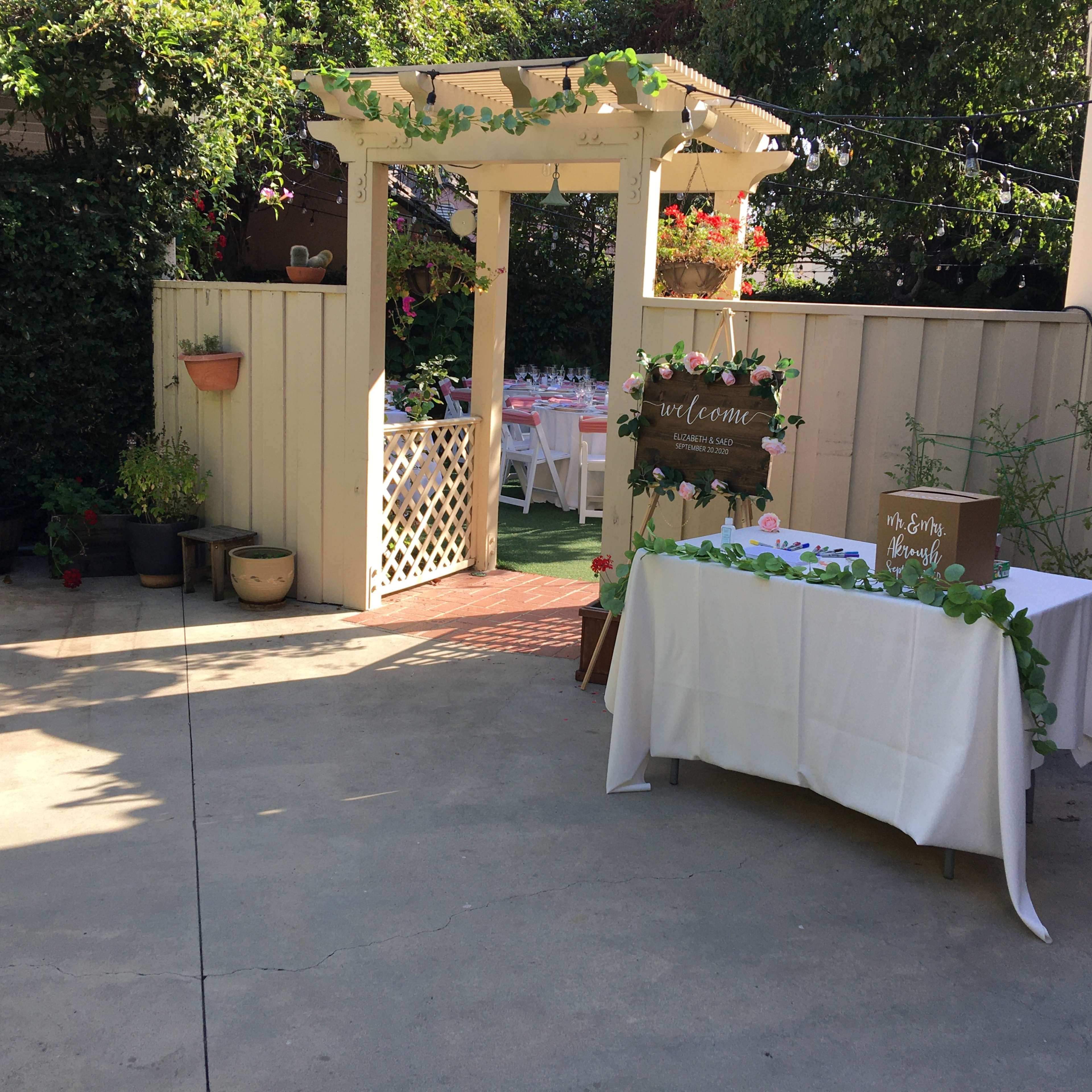 A decorated outdoor space with a white table draped in a cloth and a welcoming sign near a gazebo covered in greenery.