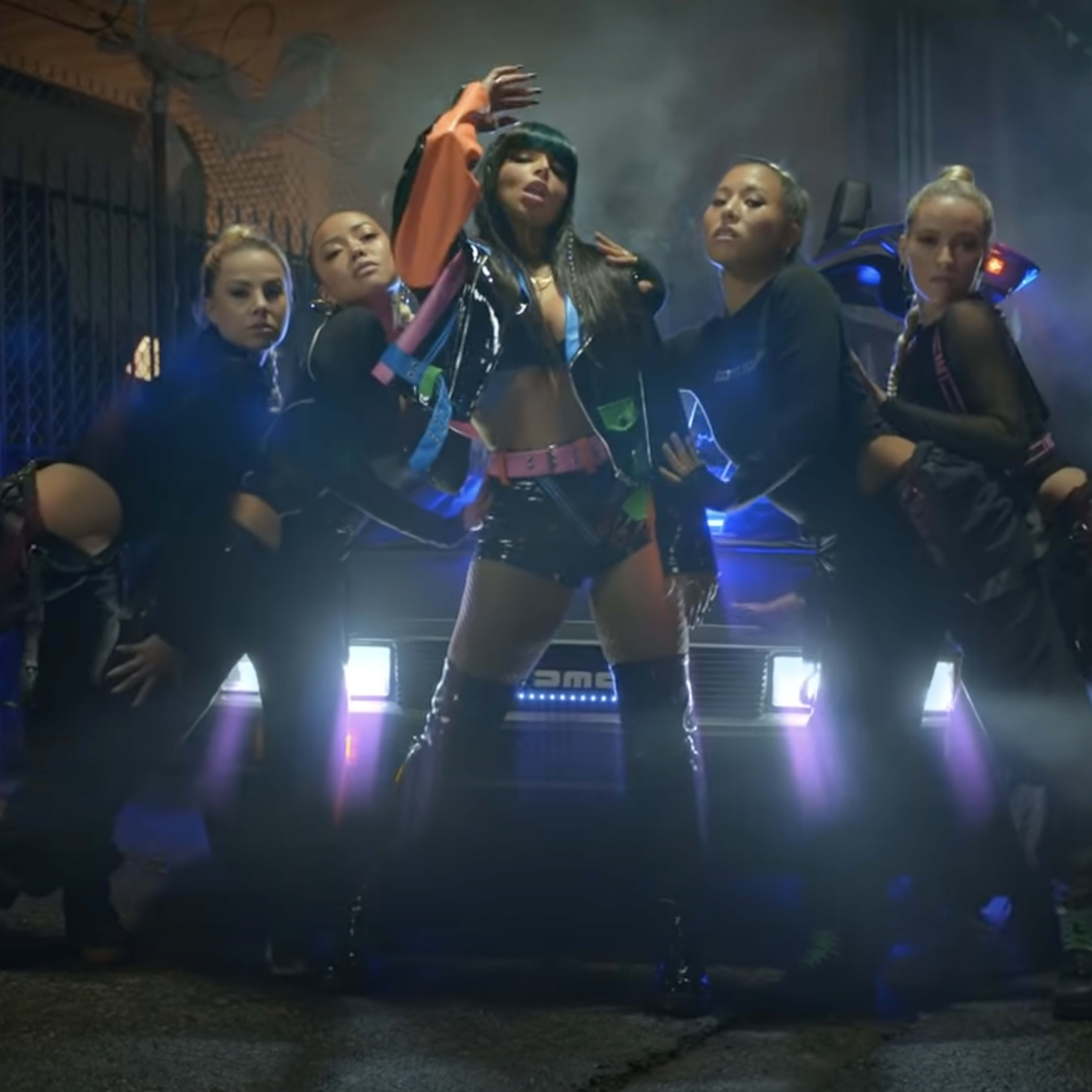 A confident performer poses at the center of a group of dancers in an urban alley, with a car illuminated by colorful lights in the background.