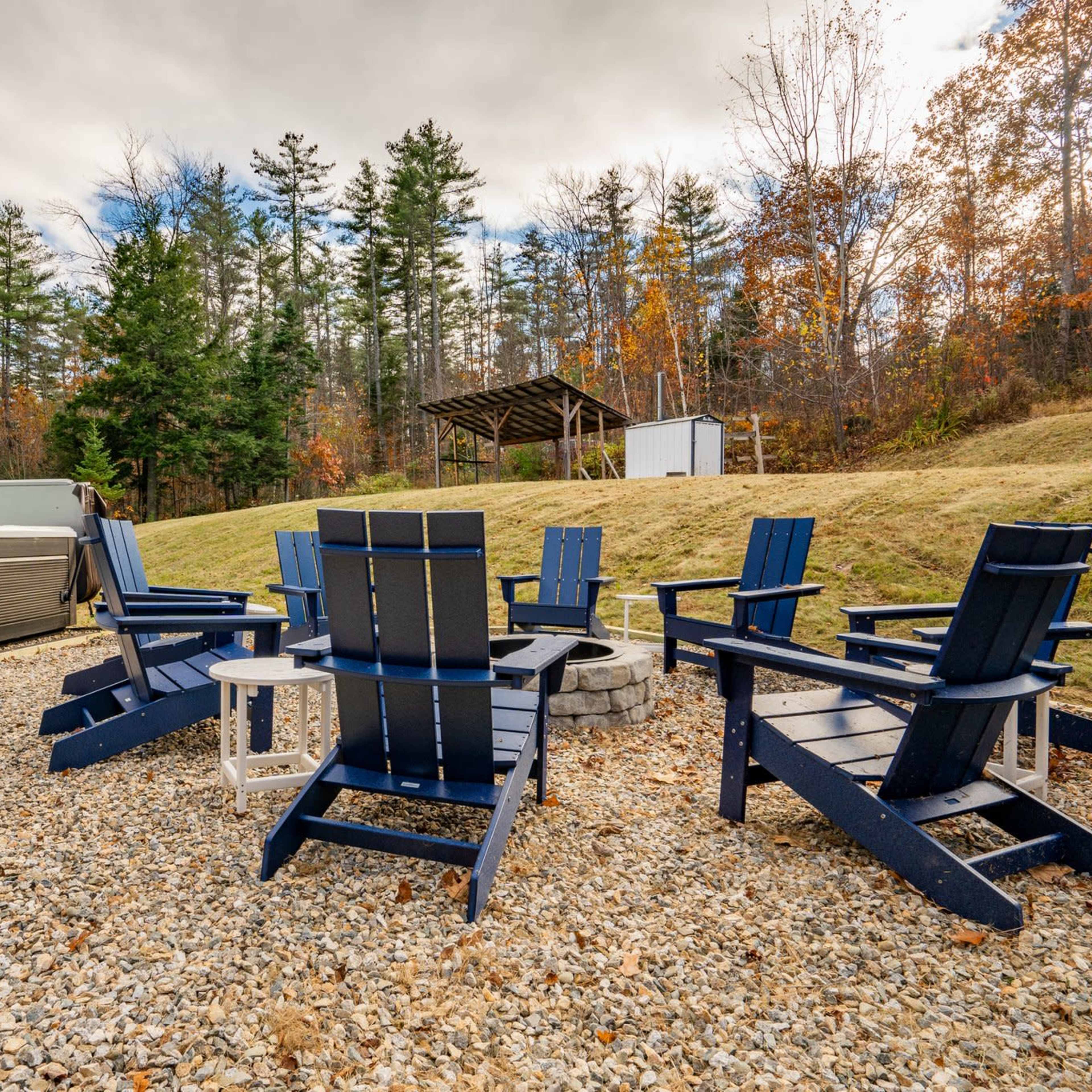 A gravel patio features several blue Adirondack chairs arranged around a fire pit, with a hot tub and wooded area in the background.