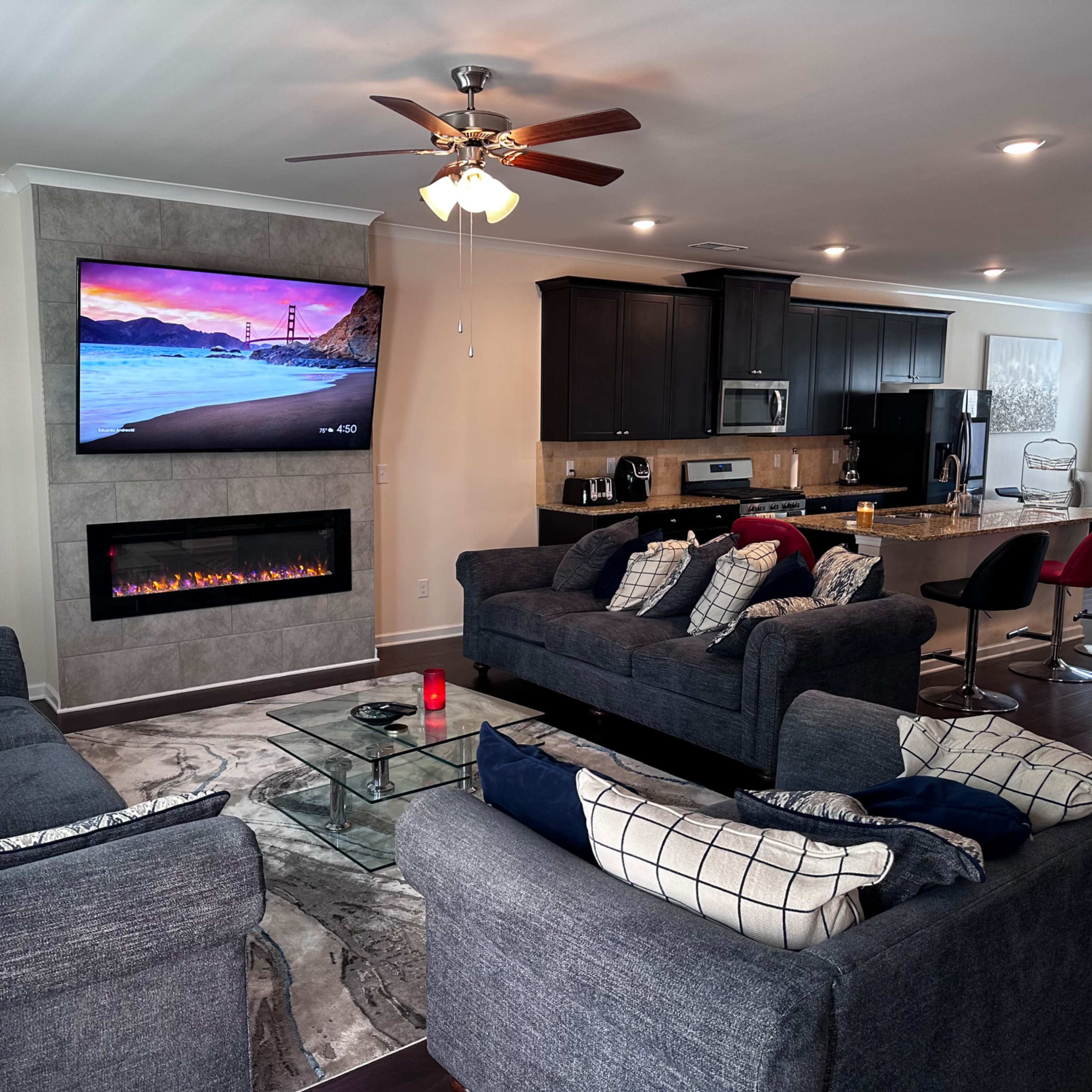 A modern living room featuring two gray sofas, a mounted television above an electric fireplace, and an open kitchen area in the background.