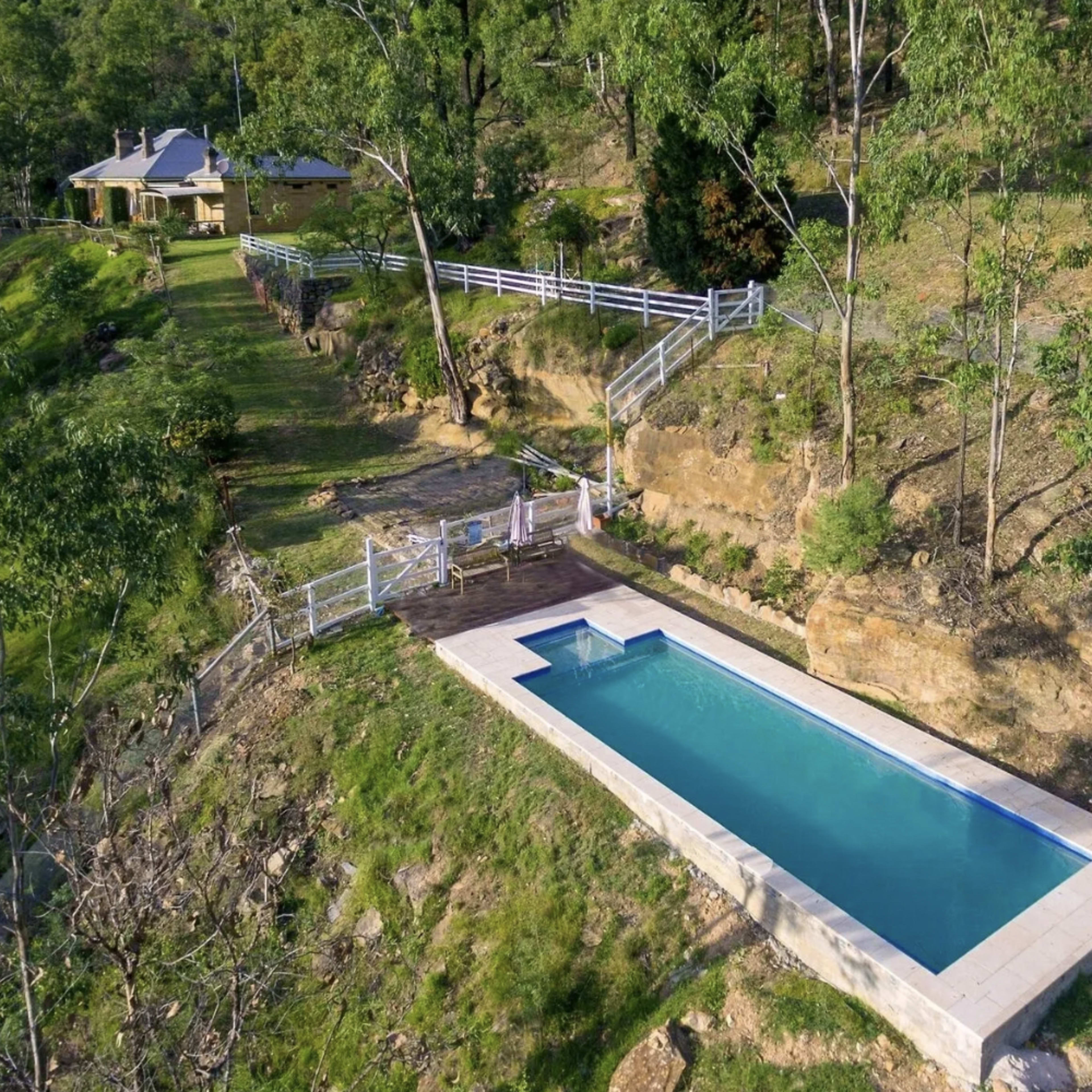 A rectangular swimming pool situated on a hillside, with a wooden deck and a house visible in the background, surrounded by greenery.