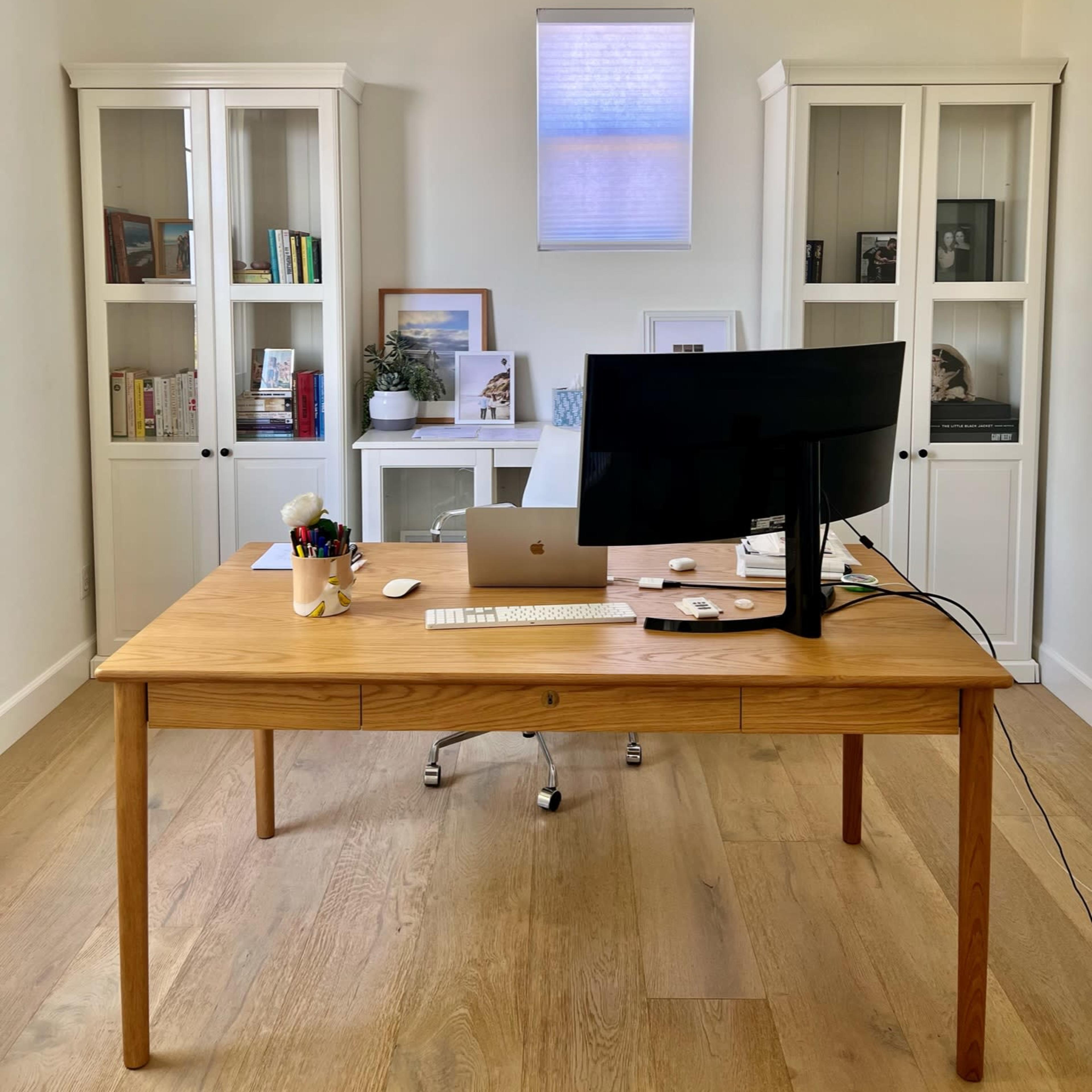 A wooden desk with a computer and office supplies is positioned in a well-lit room, flanked by two white bookshelves.