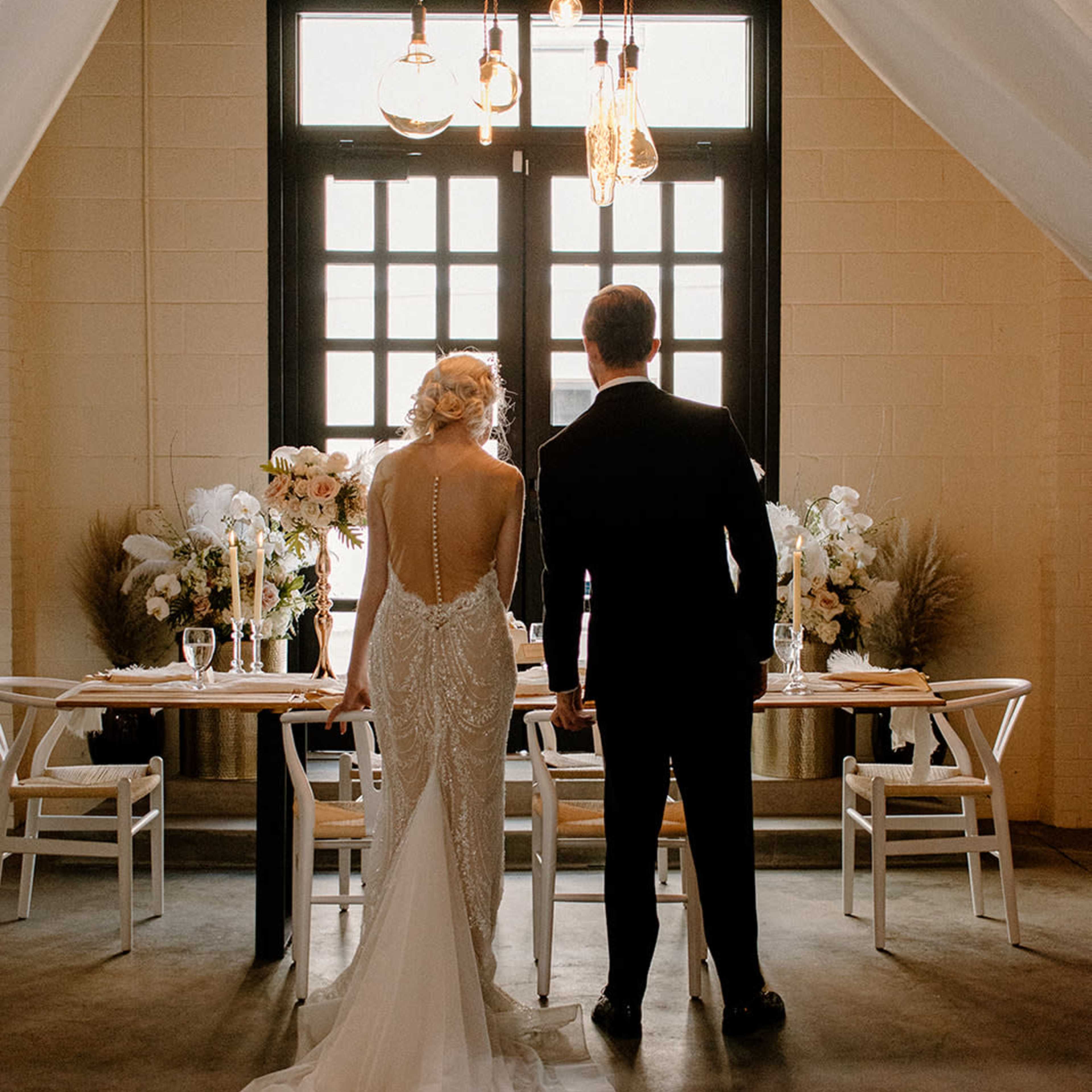 A bride and groom stand together, facing a elegantly set dining table adorned with floral arrangements and candles, under warm hanging lights in a stylish indoor venue.