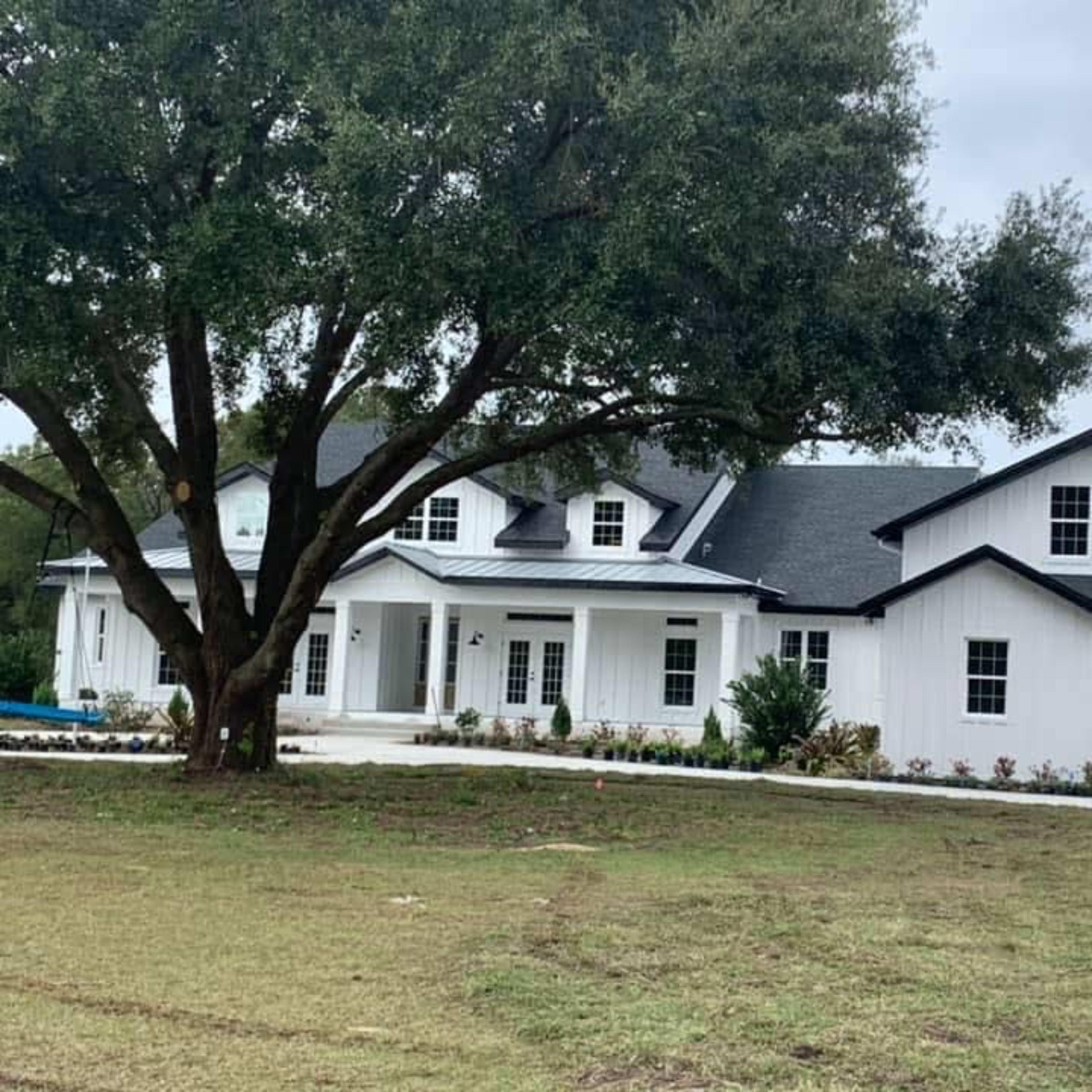 A large white house with a dark roof sits beside a grassy area and a large tree.