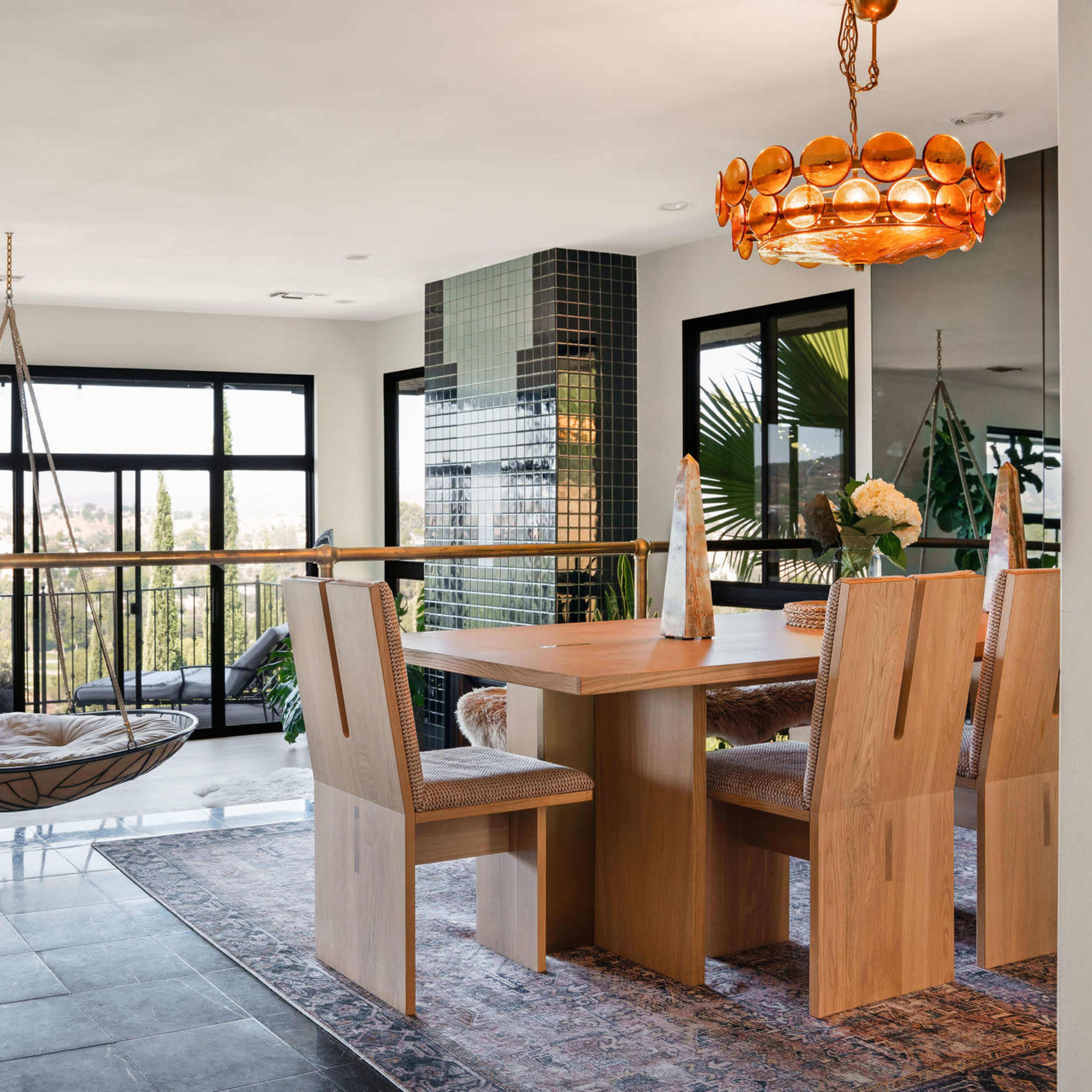 A modern dining area featuring a wooden table surrounded by light-colored chairs, a hanging chair, and large windows offering a view of the outdoors.