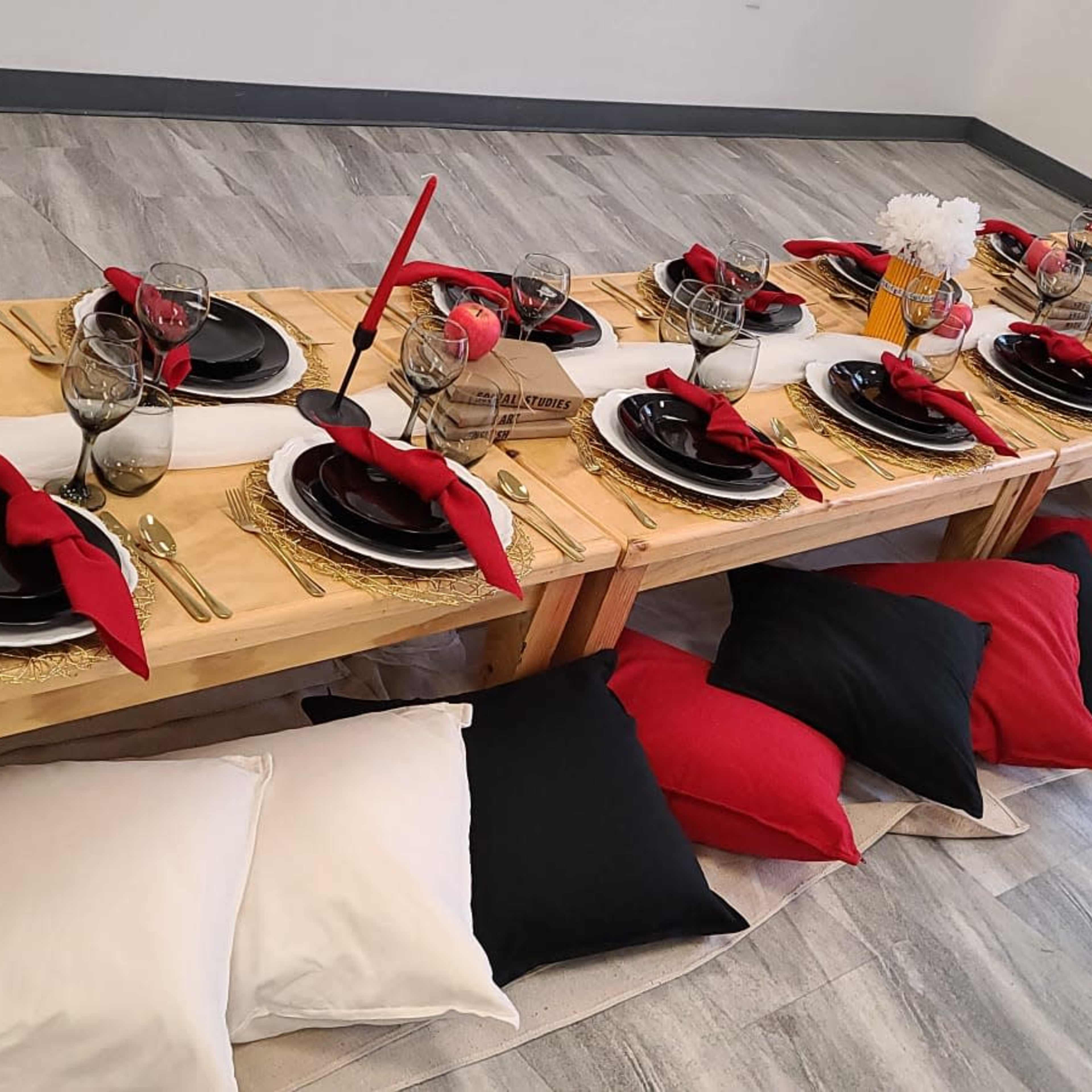 A long wooden table is set with black plates, red napkins, glassware, and red candles, surrounded by black and white cushions along the floor.