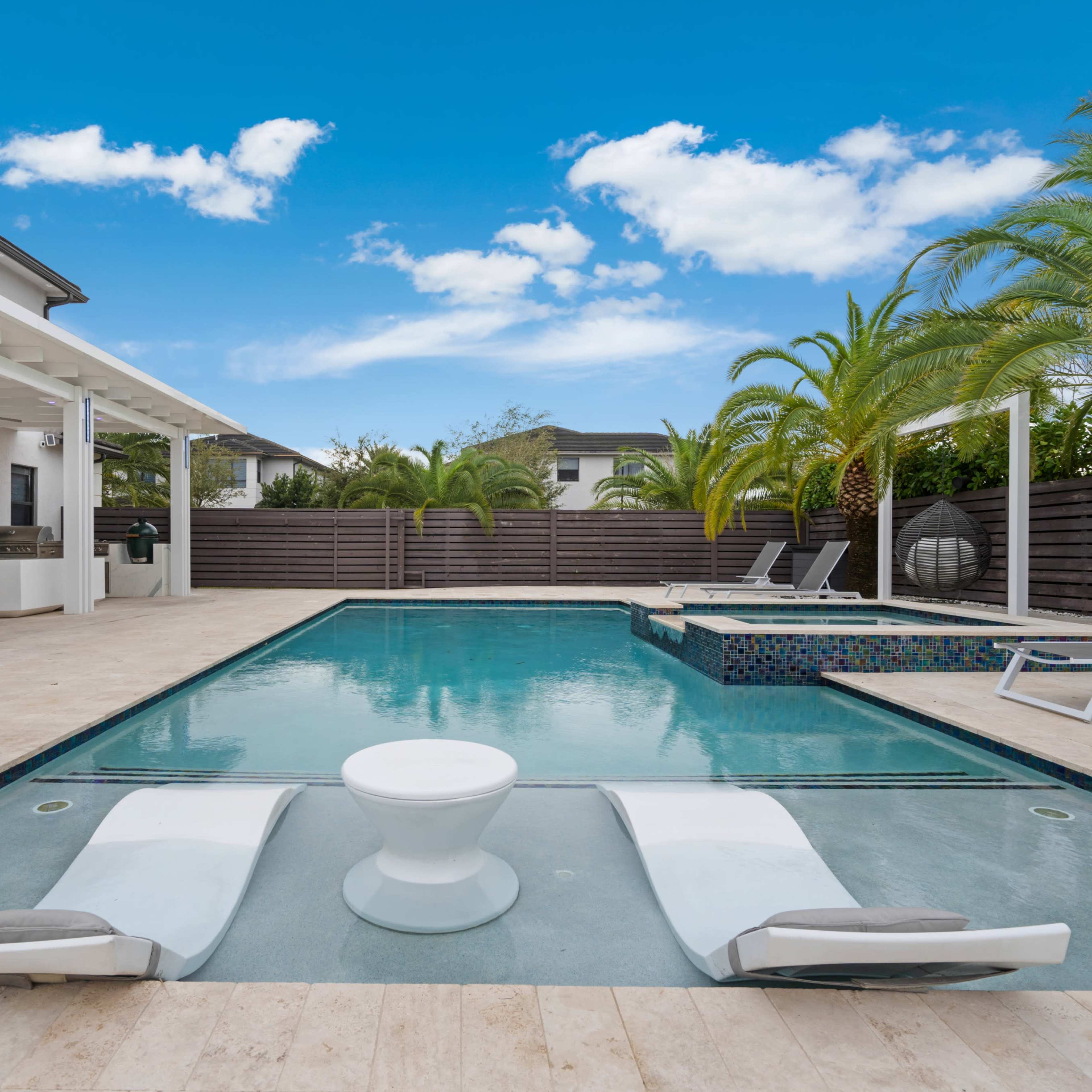 A modern backyard features a swimming pool surrounded by palm trees, lounge chairs, and a covered outdoor seating area.