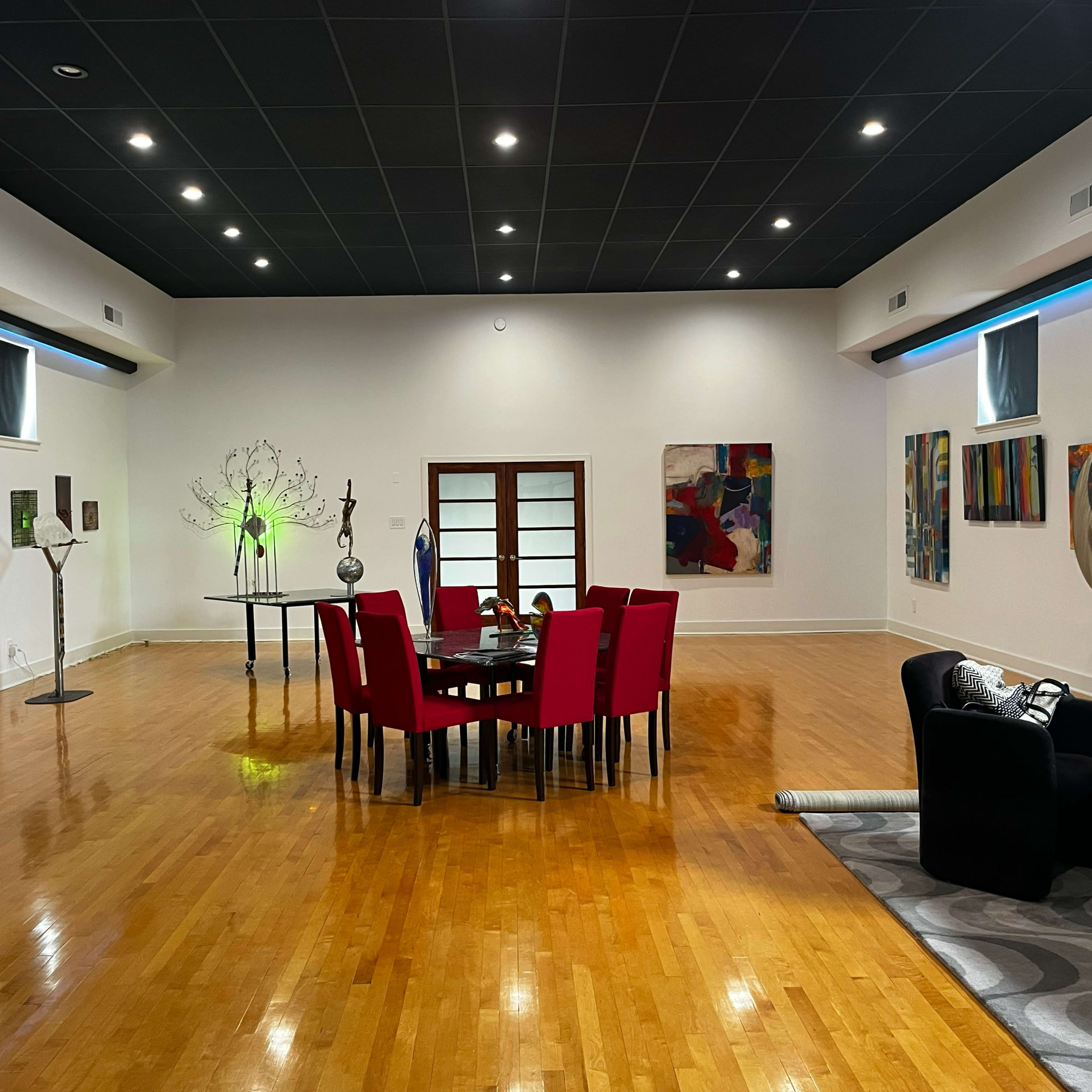 A spacious room features a round table surrounded by red chairs, with various artworks and decorative objects placed throughout.