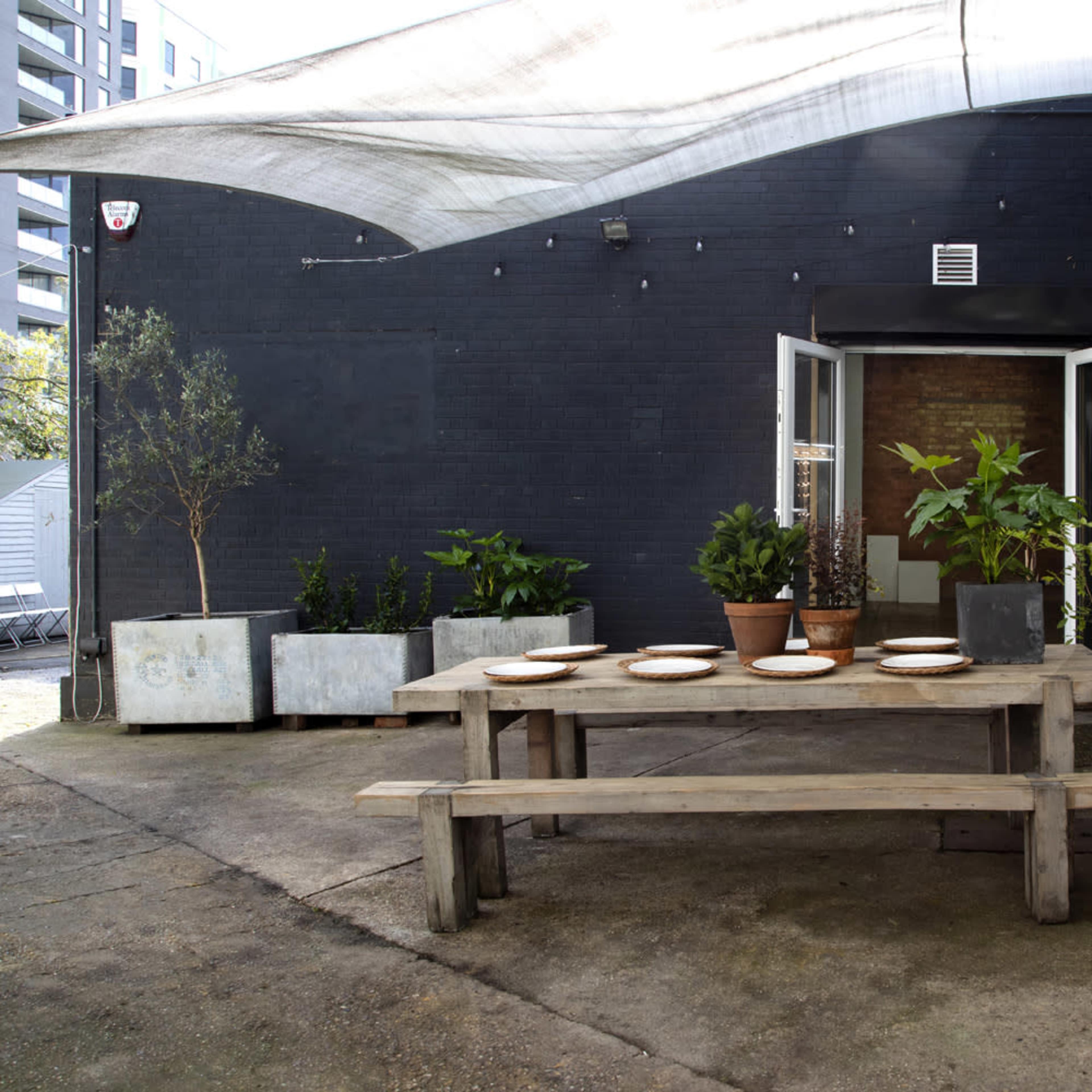 A wooden table is set with plates in a patio area surrounded by potted plants and concrete planters, with an open door leading to a dark-brick wall.