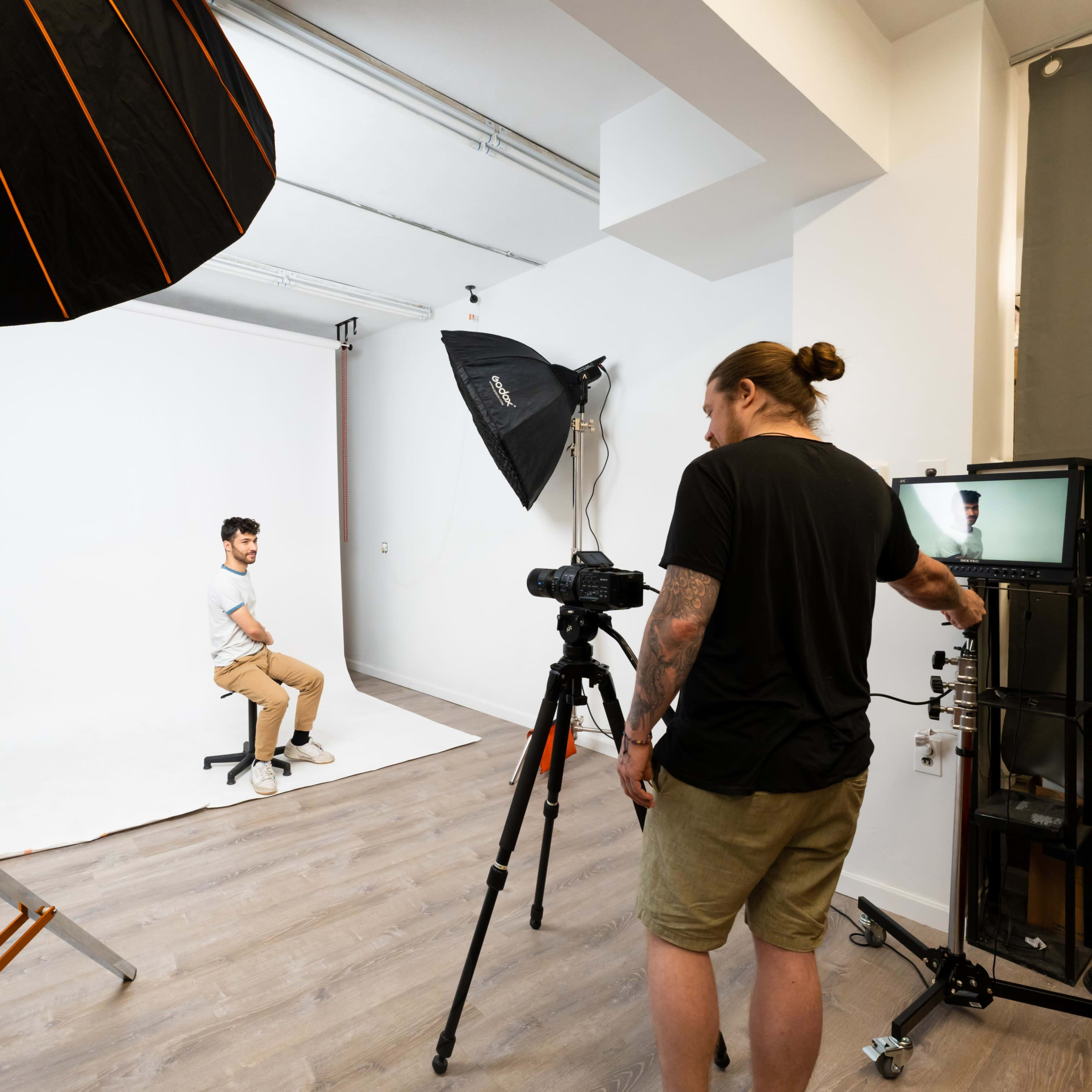 A photographer sets up a studio scene with lighting equipment and a camera while a model poses on a stool in front of a plain white backdrop.